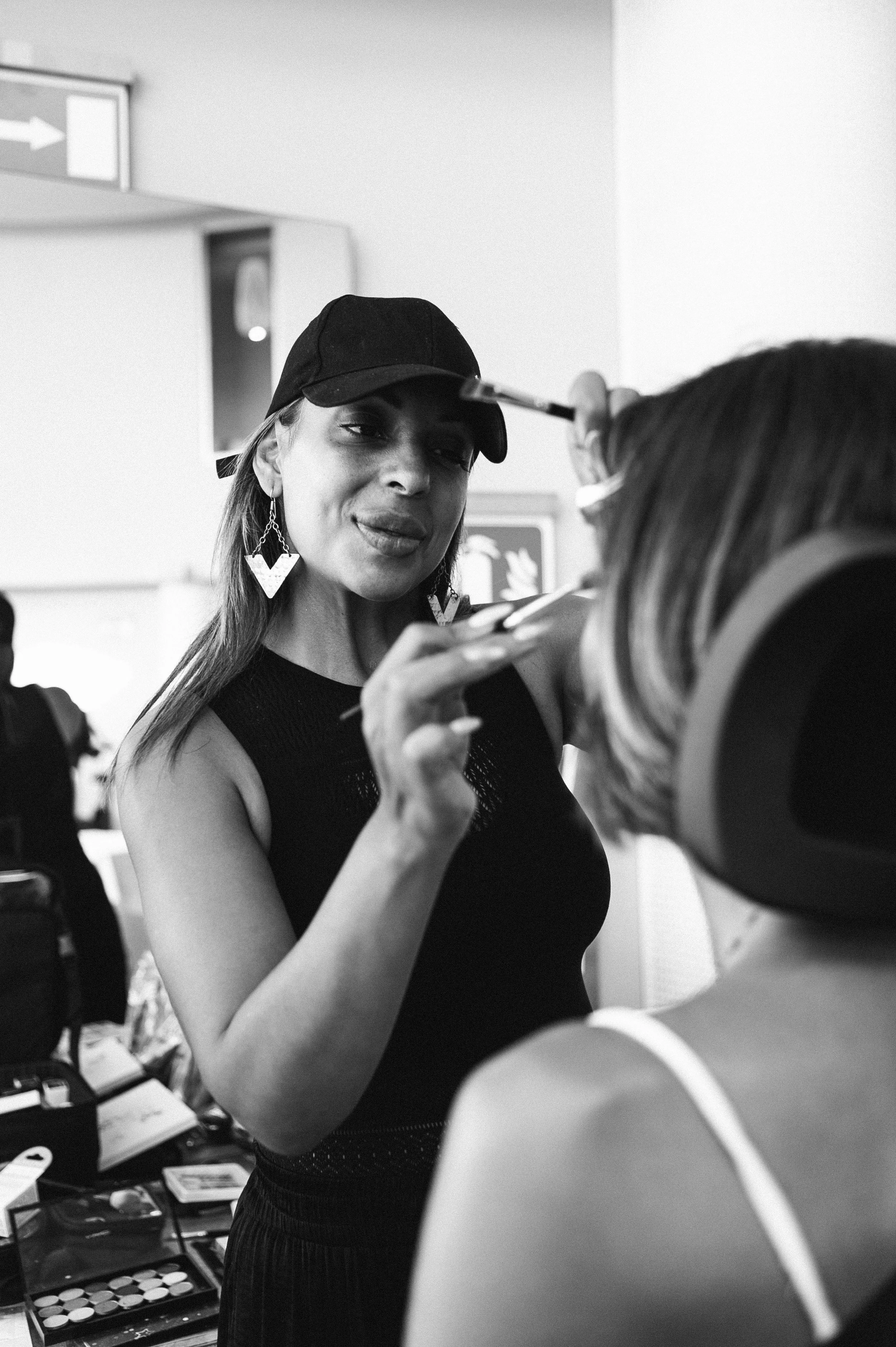 A woman with a black cap and earrings is applying makeup to another woman with striped hair at a makeup station.