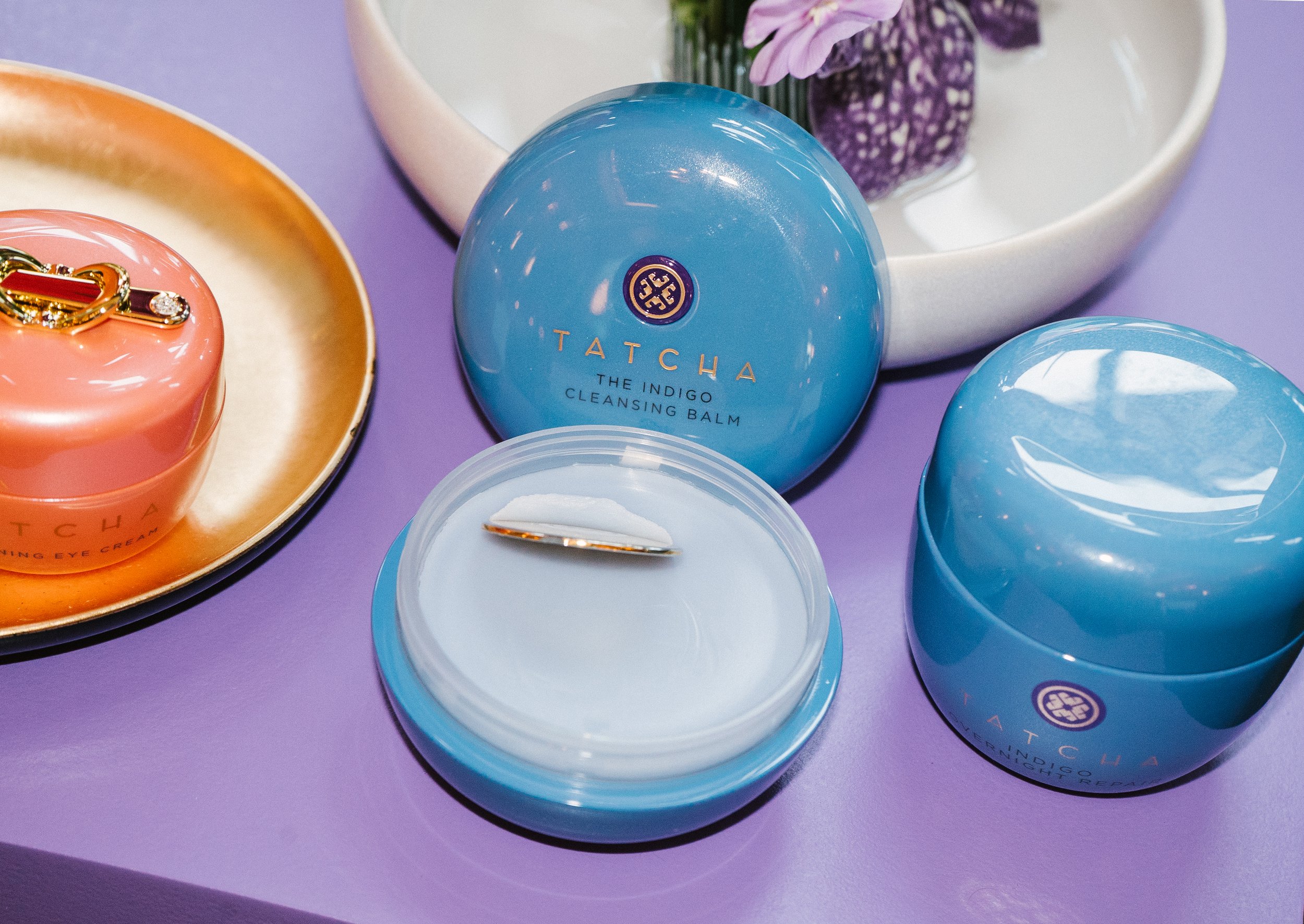 Skincare products from Tatcha, including a blue jar labeled 'Tatcha The Indigo Cleansing Balm,' an open jar with a spatula, and a pink container with jewelry, placed on a light purple surface with a white bowl containing purple and green flowers.