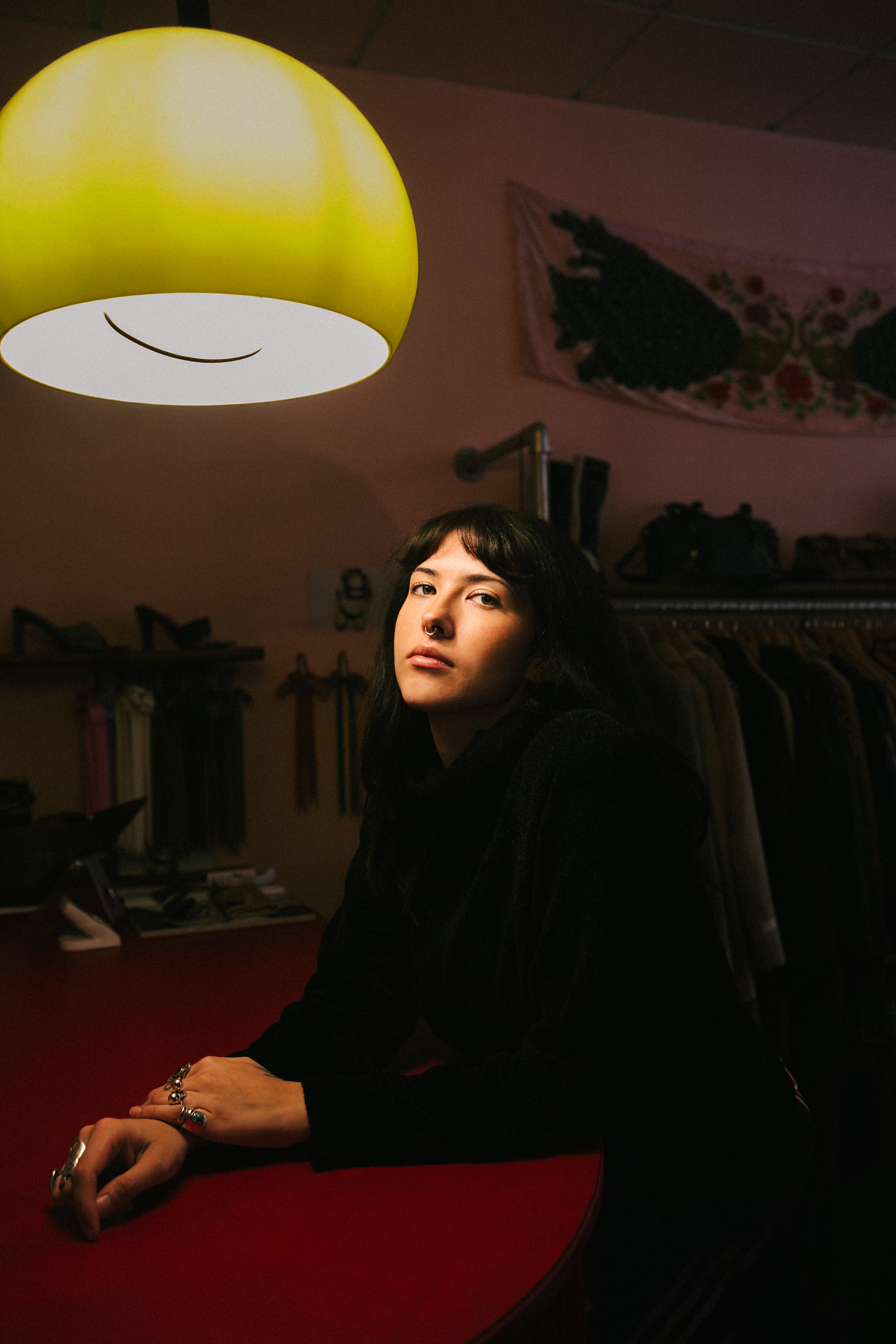 A woman with dark hair and multiple rings resting her hand on a red table in a dimly lit room, with a yellow lampshade hanging above her.