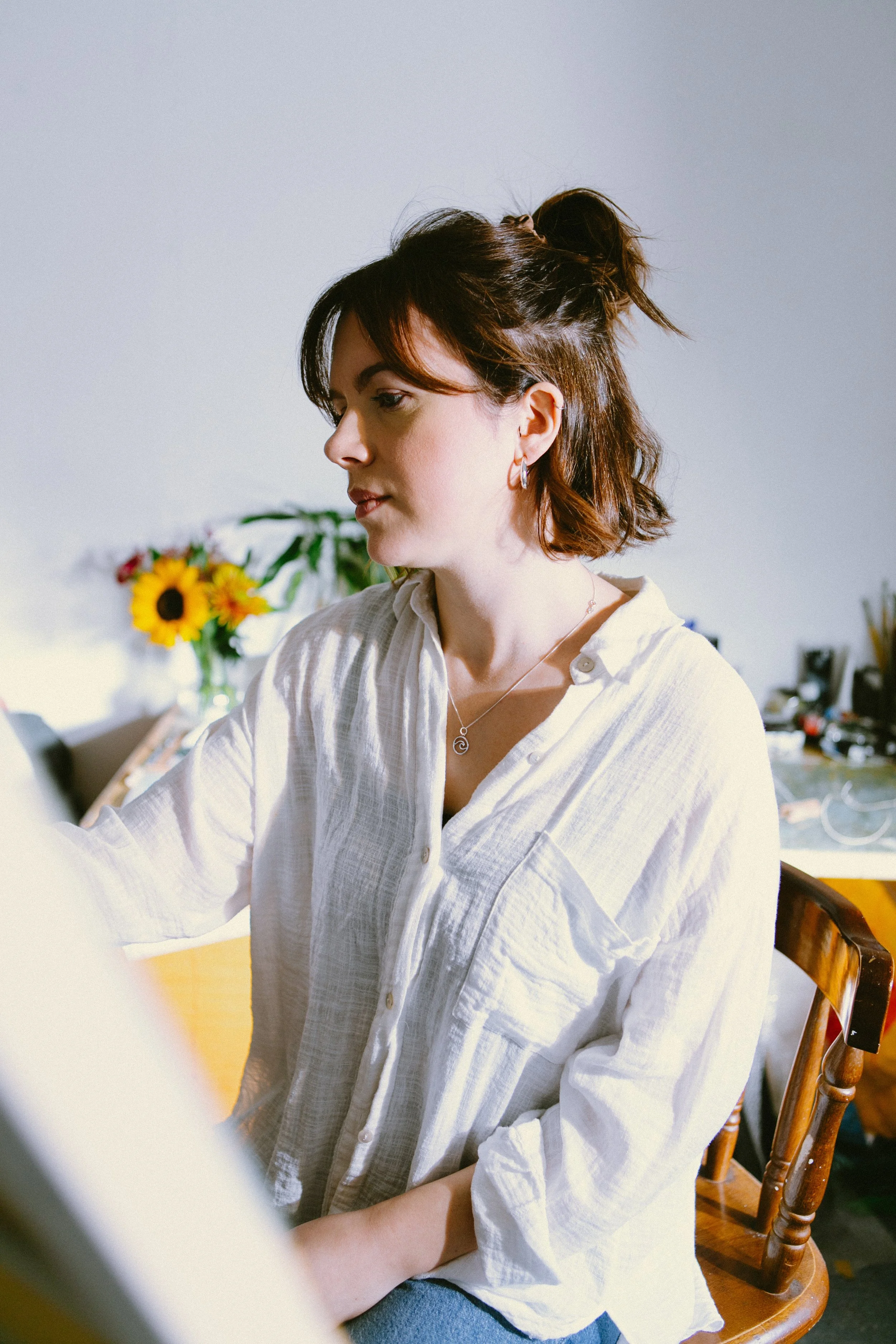 A woman with short, brown hair styled in a half-up bun, wearing a white button-up shirt and silver jewelry, sitting in a room with a sunflower in a vase and a desk with various items in the background.
