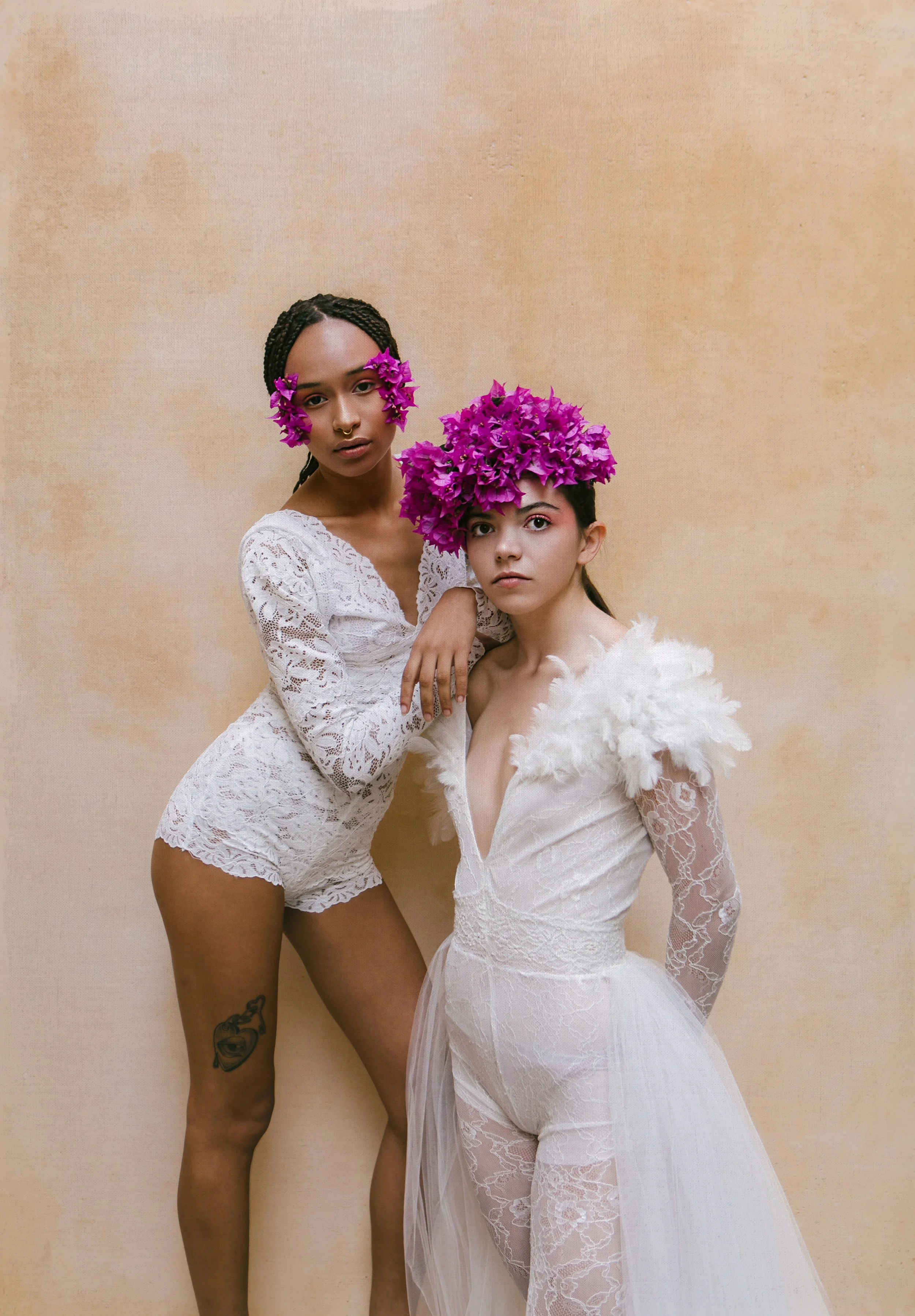 Two women in white attire with purple flower crowns pose against a beige background. One woman stands tall in lace shorts and the other sits in a lace dress with feathered shoulders.