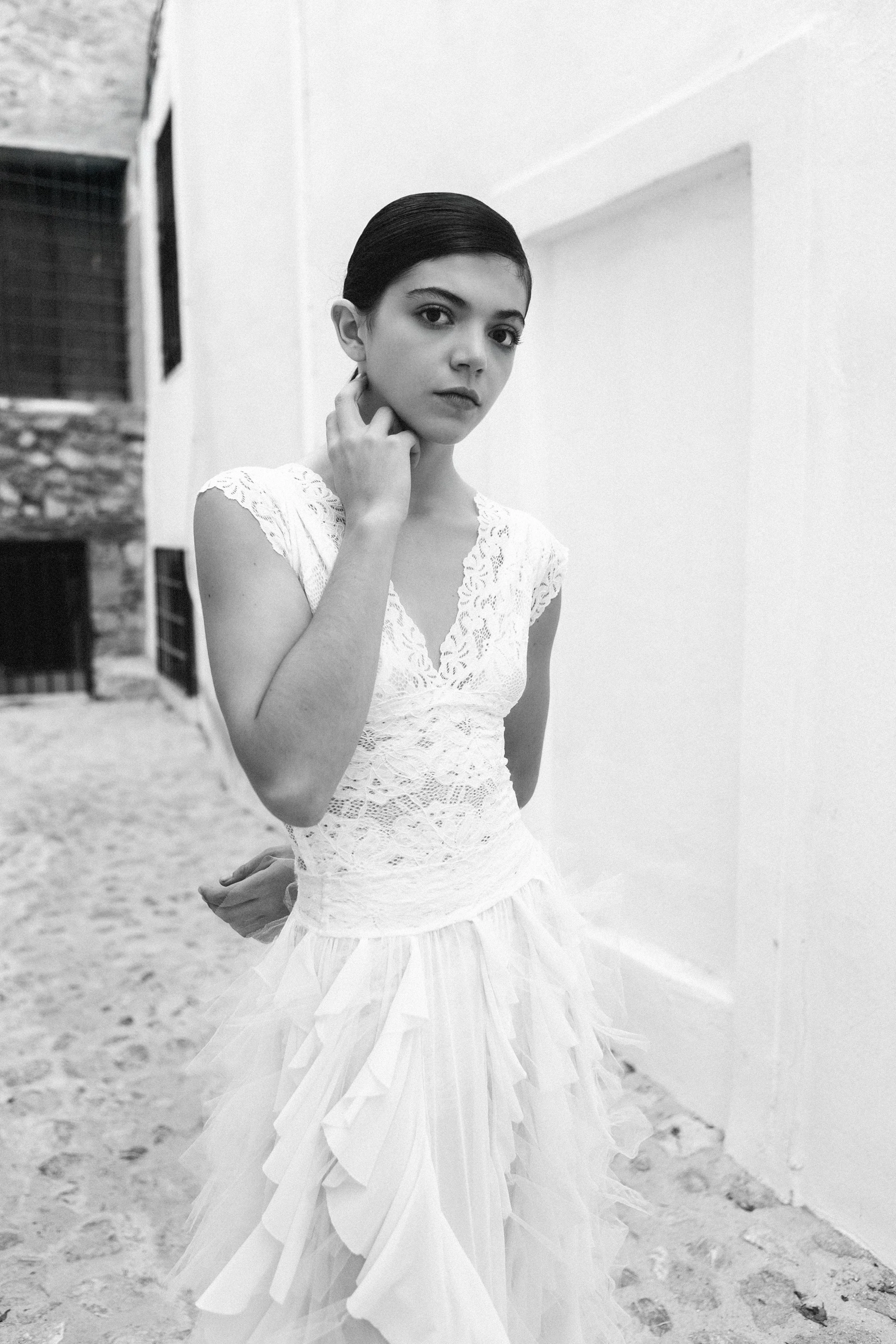 A young woman in a white lace dress standing outdoors on a stone-paved ground, with a white wall to her right and an old building with barred windows to her left.