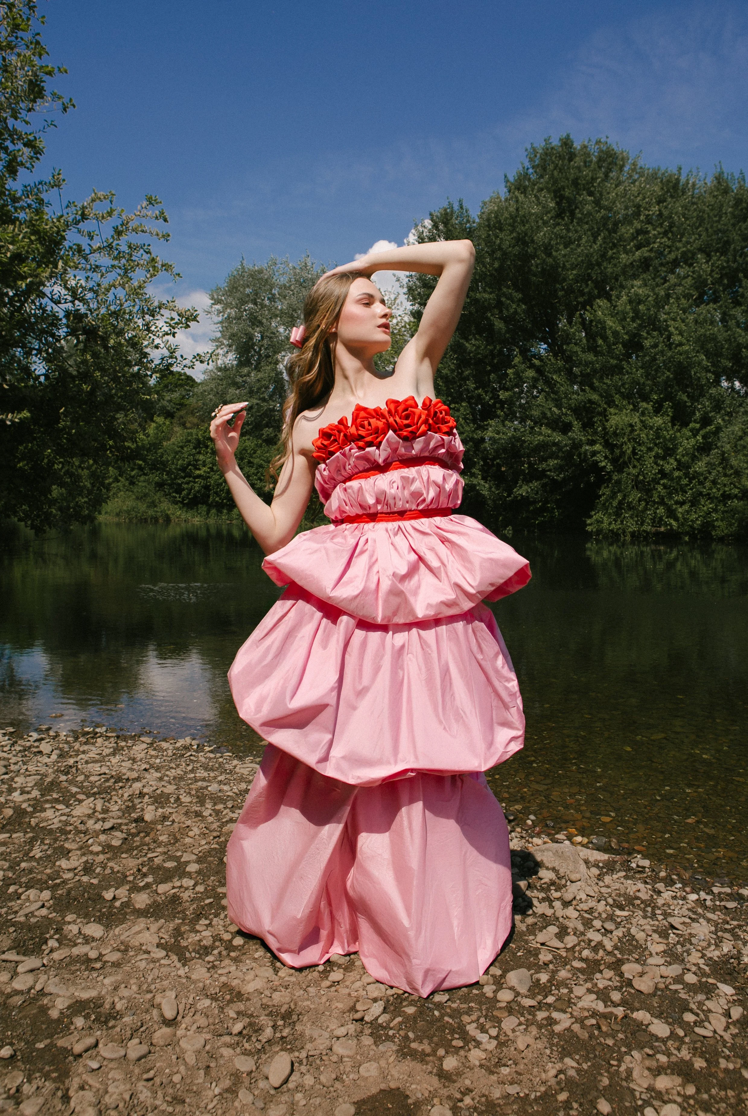 A woman in a pink, elaborate dress with ruffles and floral accents standing on a rocky shoreline by a body of water, with trees and a blue sky in the background.
