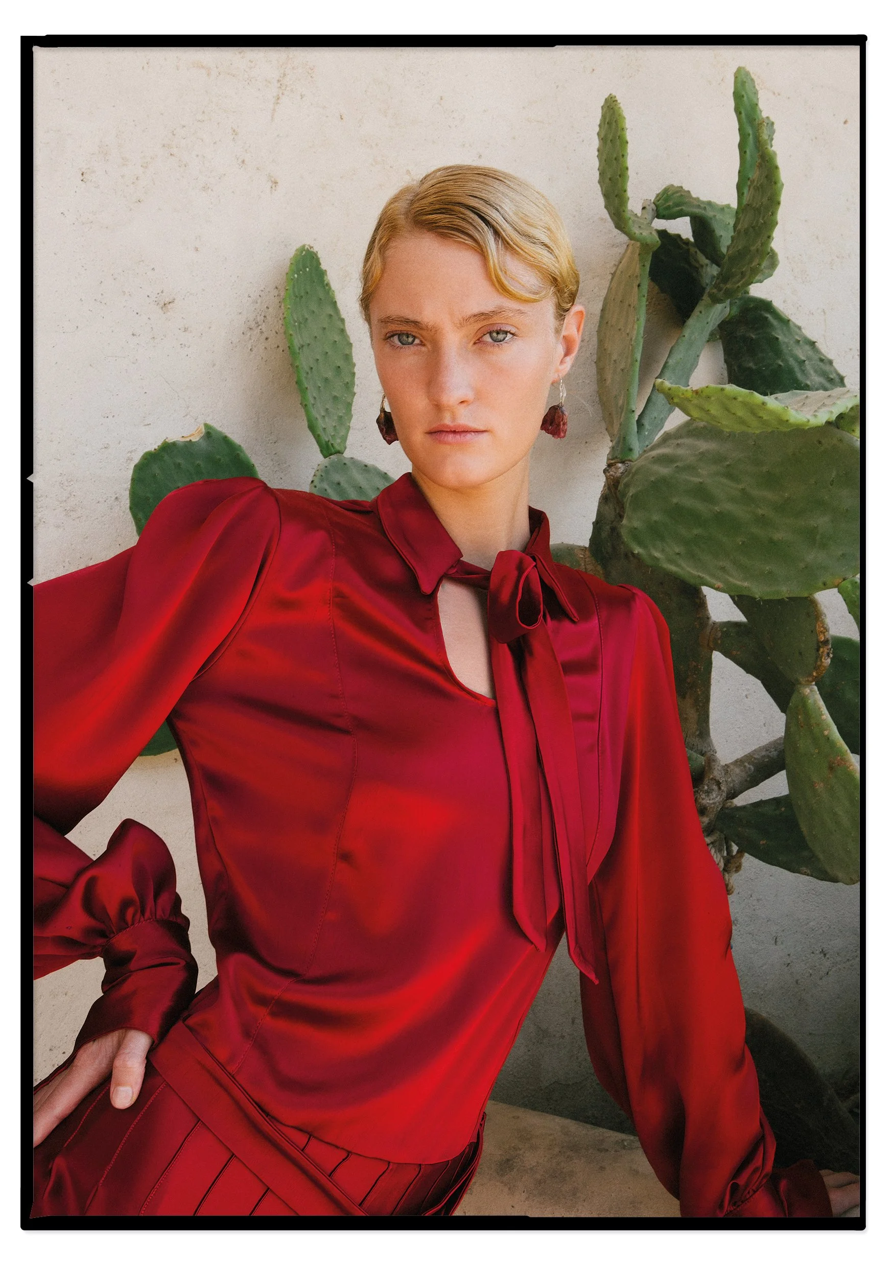 A woman with short blonde hair wearing a red satin blouse with a bow tie at the collar, standing in front of a cactus plant against a light-colored wall.