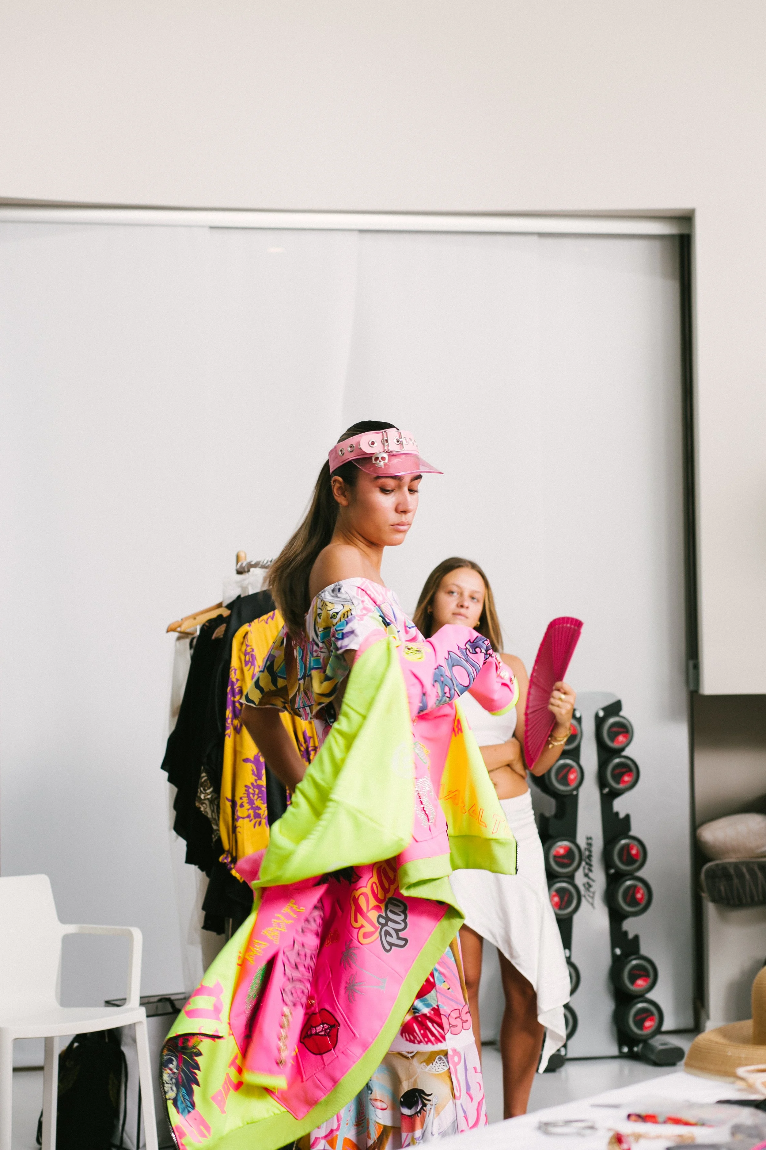 Two women in colorful clothing in a room with a rack of hanging clothes and a storage unit with black and red objects in the background.