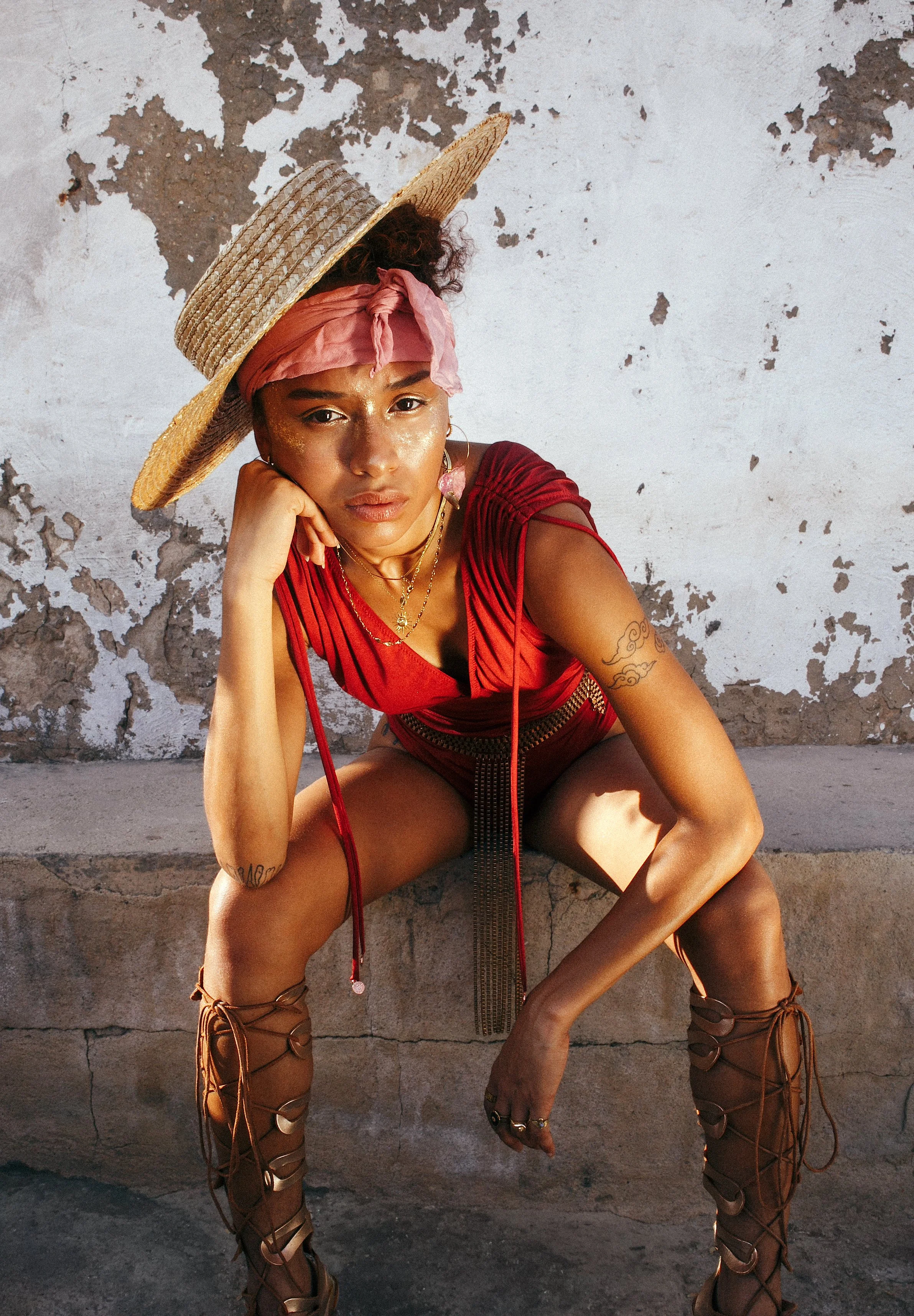 A woman wearing a large straw hat, red top, and gladiator sandals straddles a concrete ledge, sitting against a weathered, peeling wall.