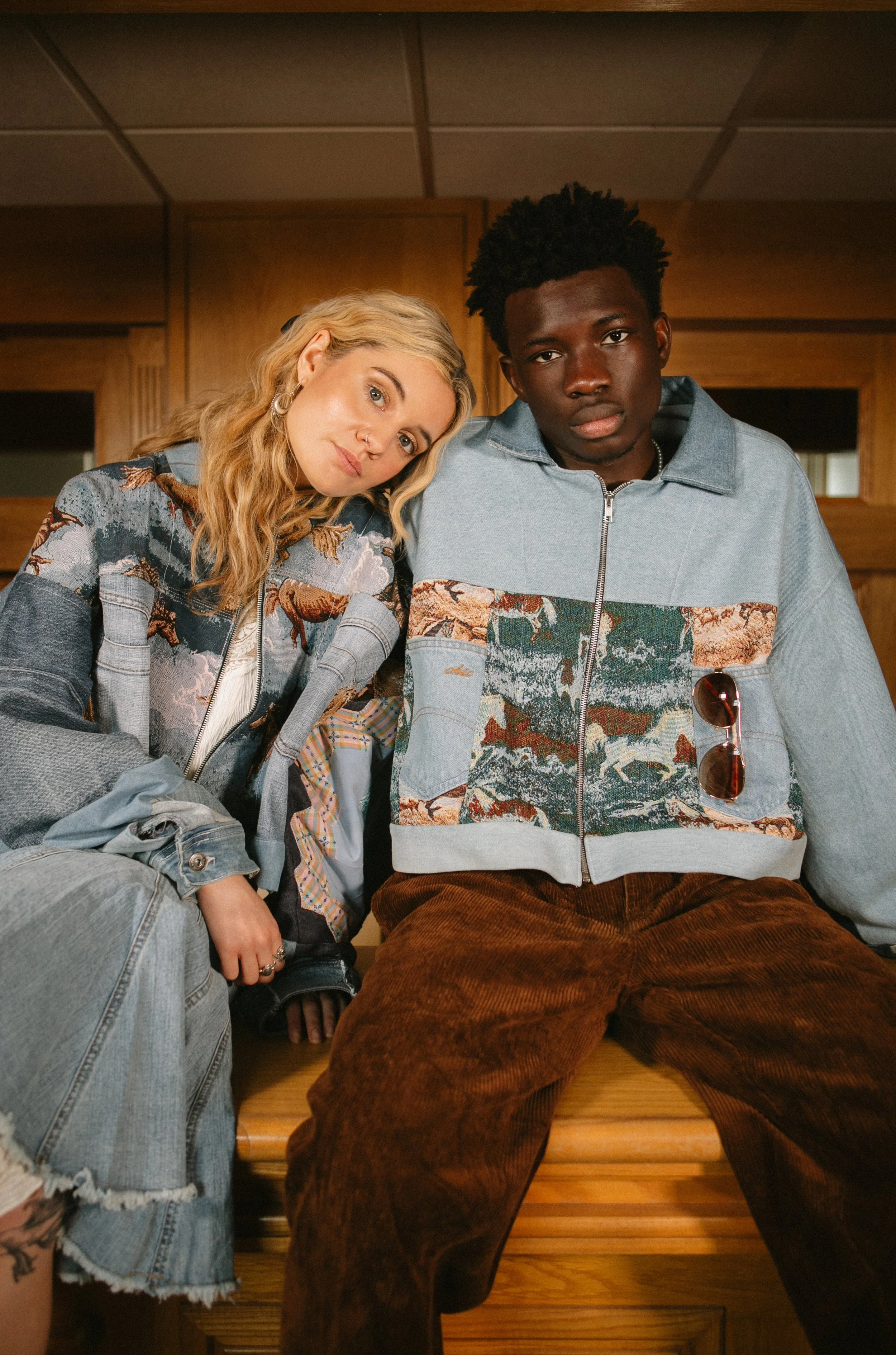Two young adults sitting close together indoors, wearing colorful jackets with nature-themed patterns, looking at the camera with neutral expressions.