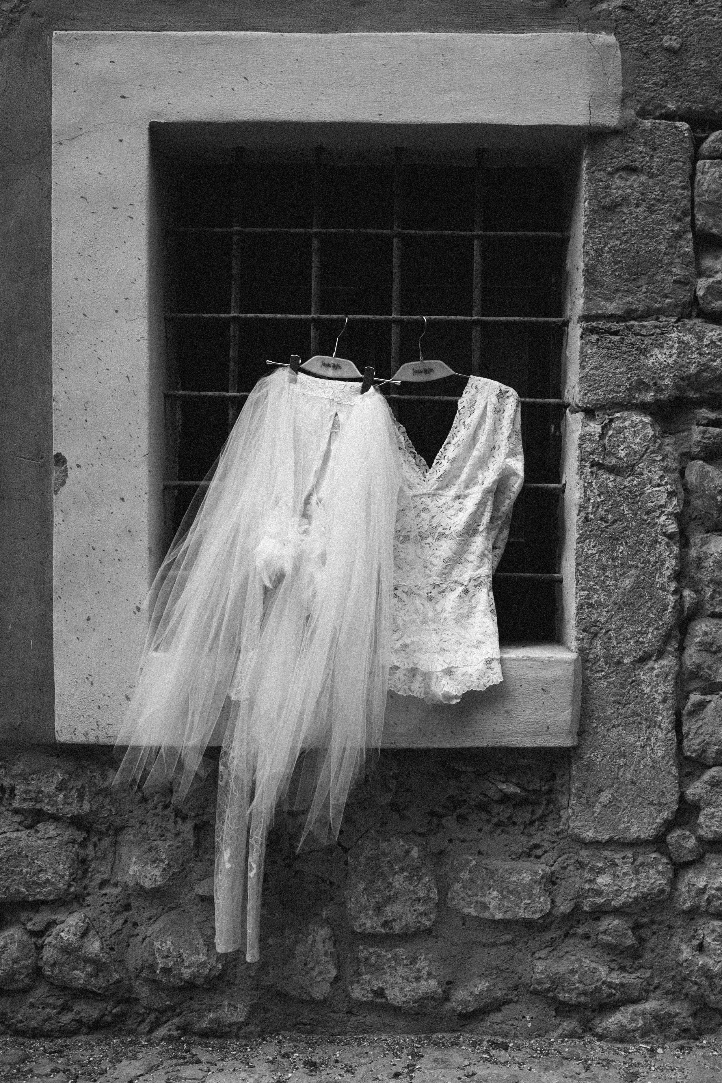 A pair of white bridal clothes, a tulle skirt and a lace top, hanging on hangers in a window with a stone and brick wall background.
