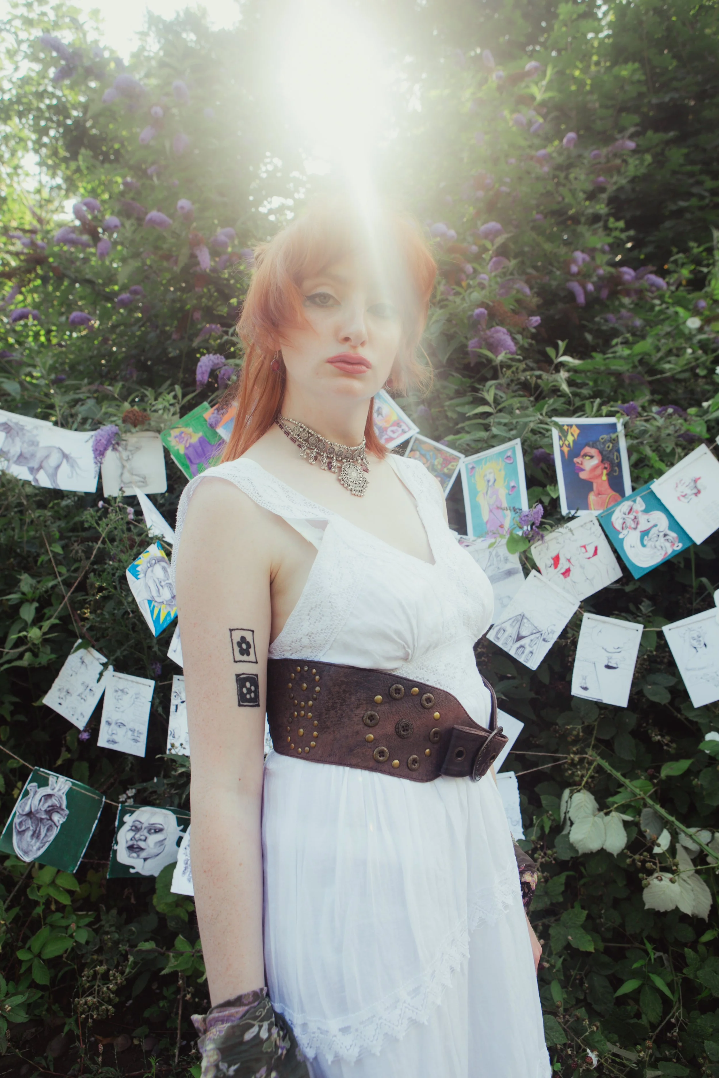 A woman with red hair in a white dress and decorative jewelry stands outdoors, with artwork hanging behind her on a string among green foliage and purple flowers.