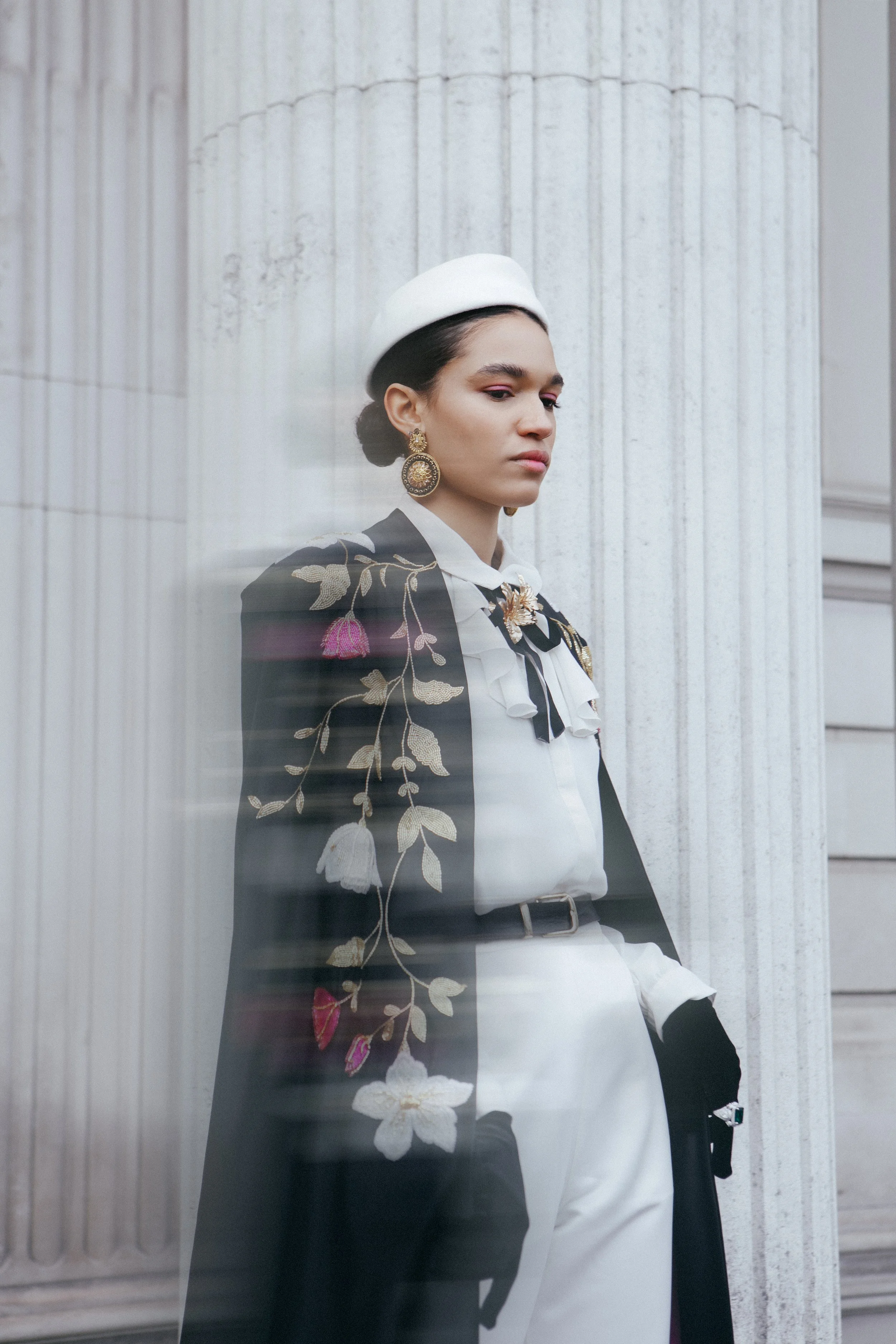 A woman dressed in a stylish black and white outfit with floral embroidery, gold earrings, and a white hat, standing against a classical columned building.
