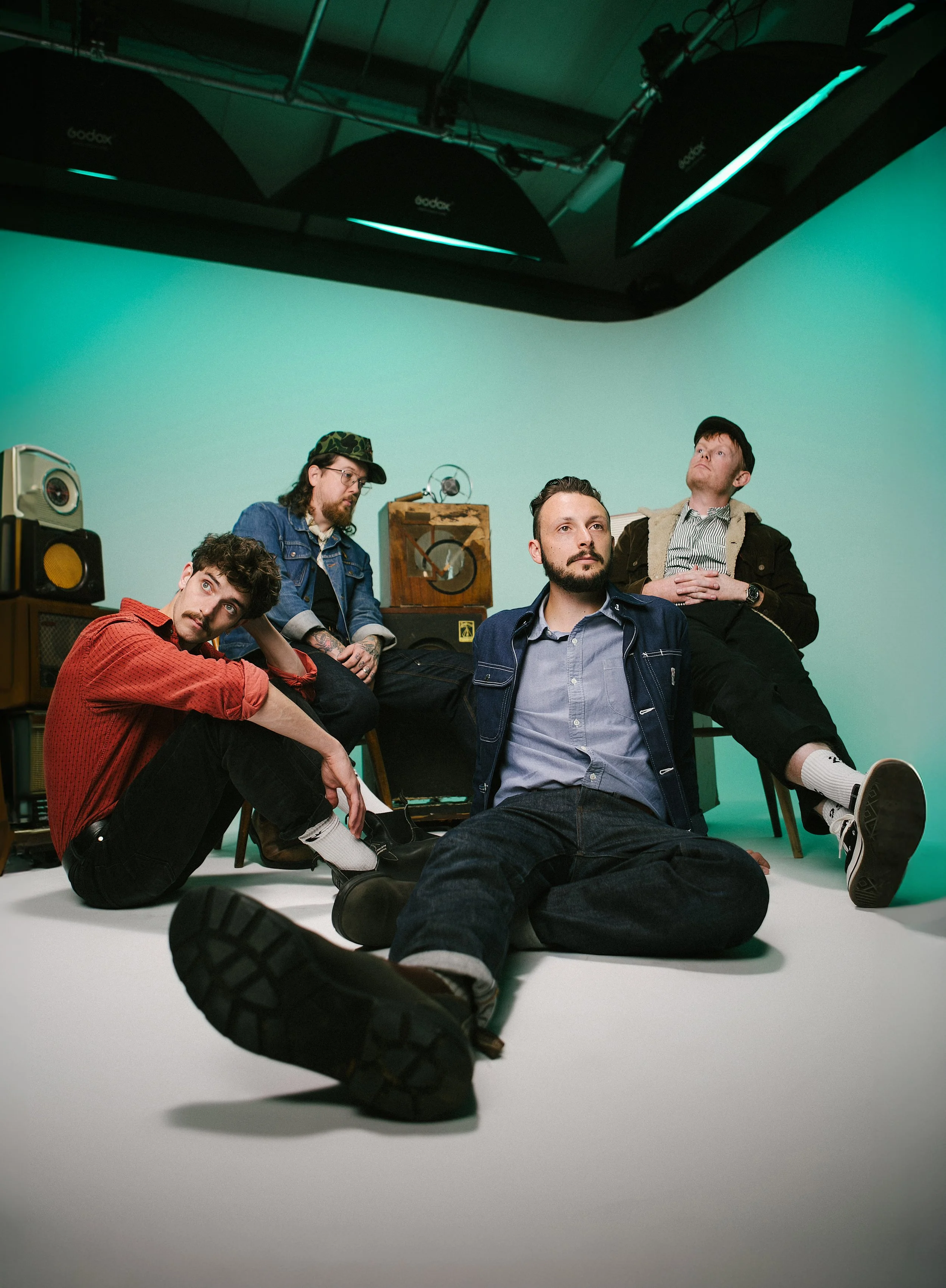 Four men in a photo studio with a green screen background. They are sitting and reclining on the floor and furniture, with vintage speakers and equipment behind them.