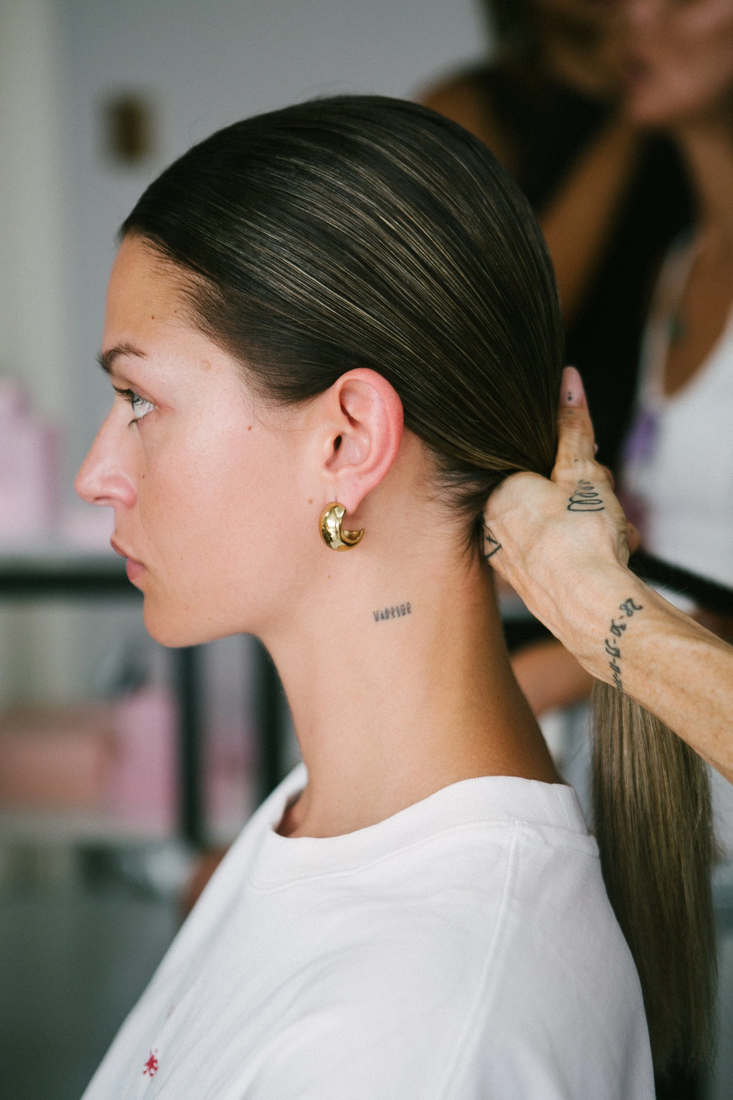 A woman with dark, slicked-back hair and gold hoop earrings has her hair styled by a tattooed person with a tattooed hand, while sitting in a space that appears to be a salon or similar setting.