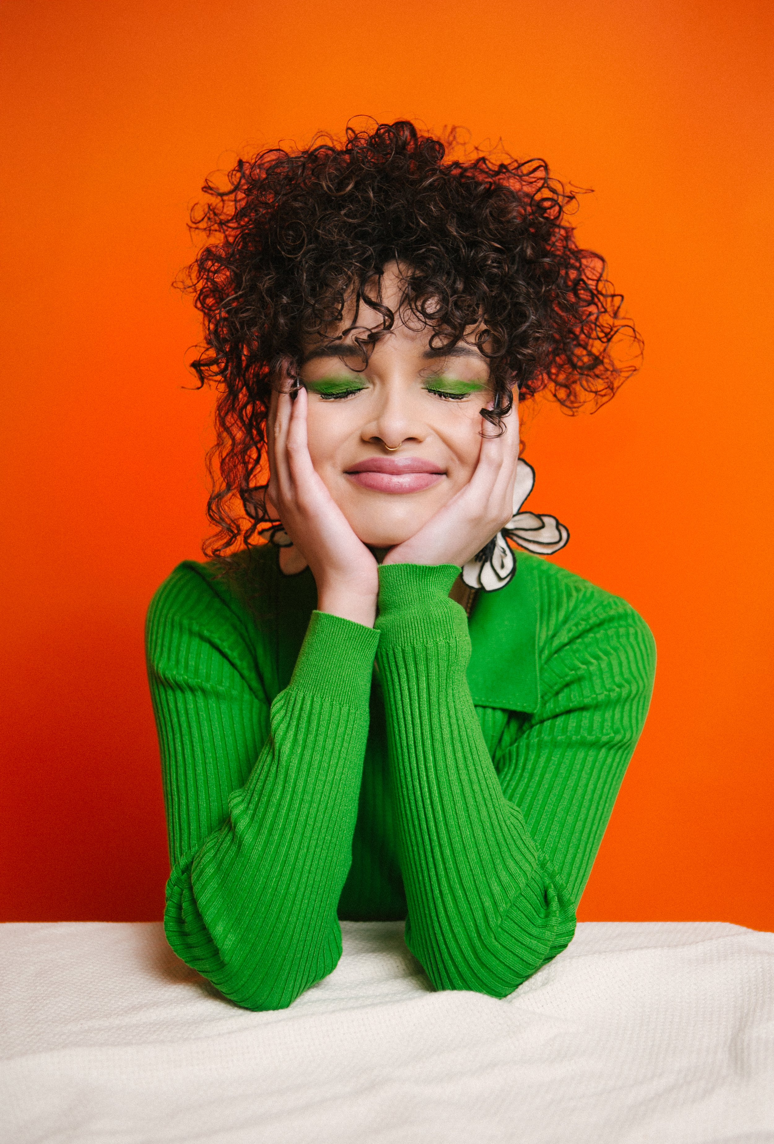 A young woman with curly dark hair and green eye makeup resting her face on her hands, wearing a bright green long-sleeved top, with a vibrant orange background.
