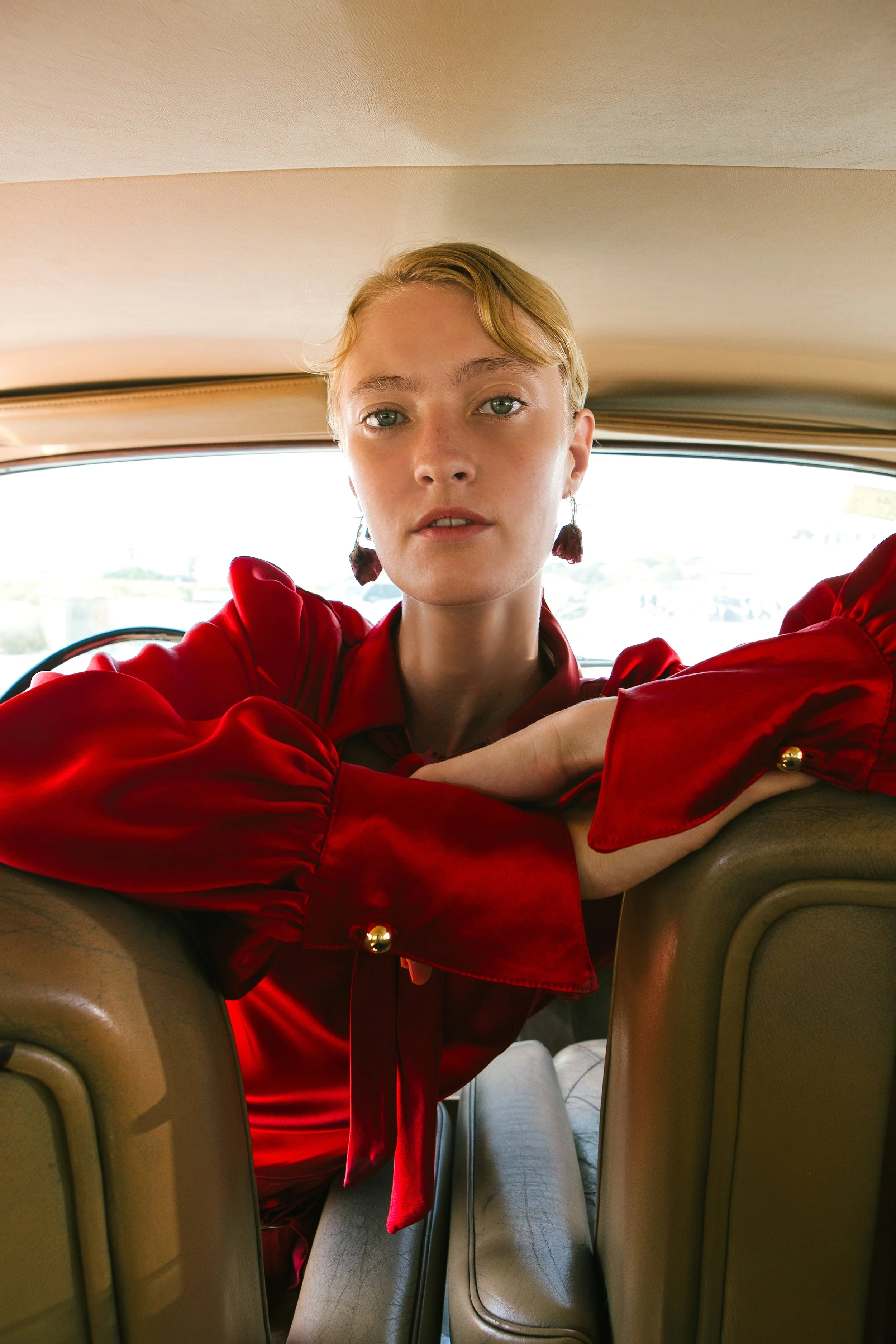 A woman with short blonde hair sitting in a vintage car, wearing a red satin blouse with puffy sleeves and gold button details, looking directly at the camera.