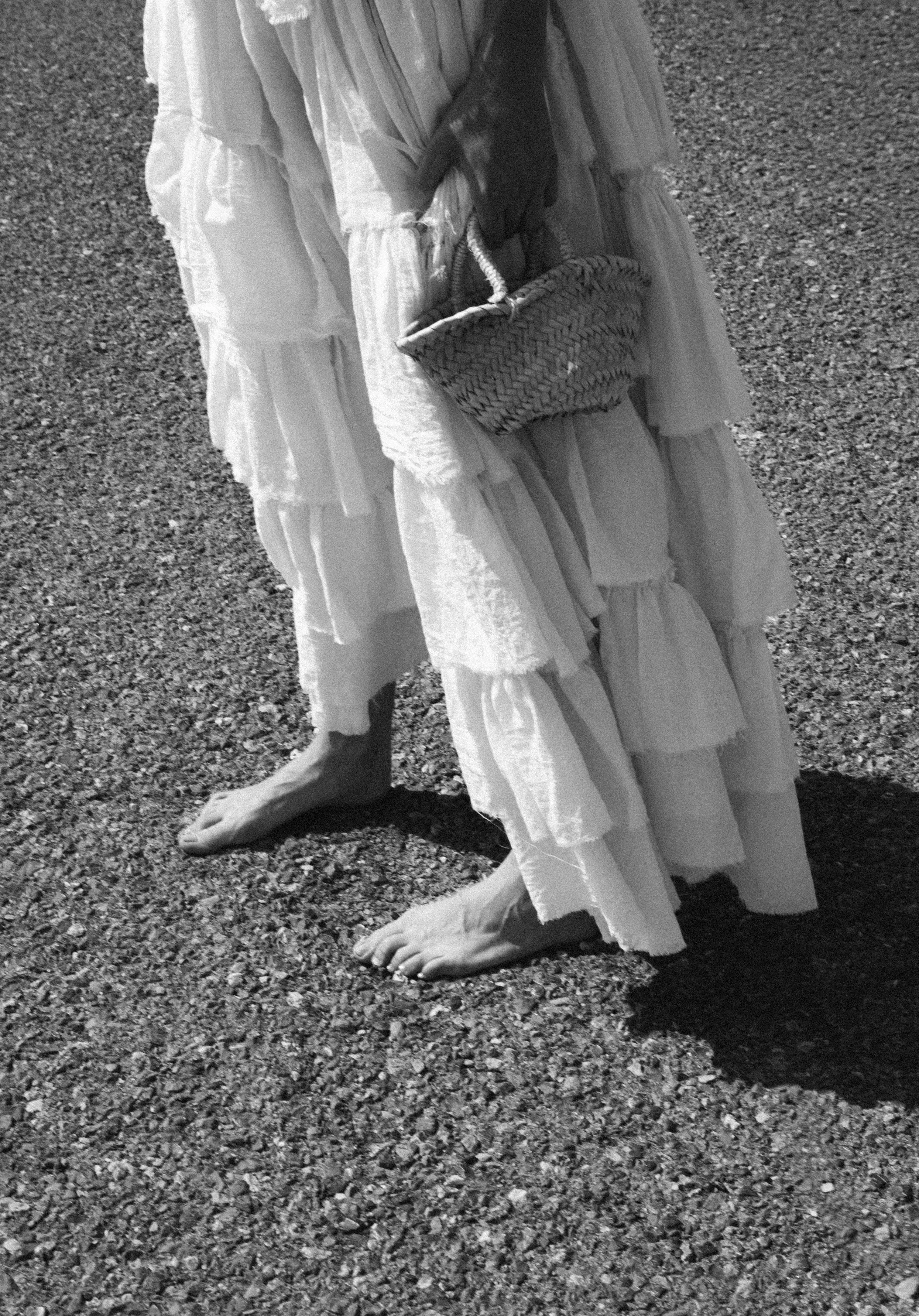 A person standing barefoot on gravel ground, wearing a long, flowing, tiered white dress, holding a woven bag.