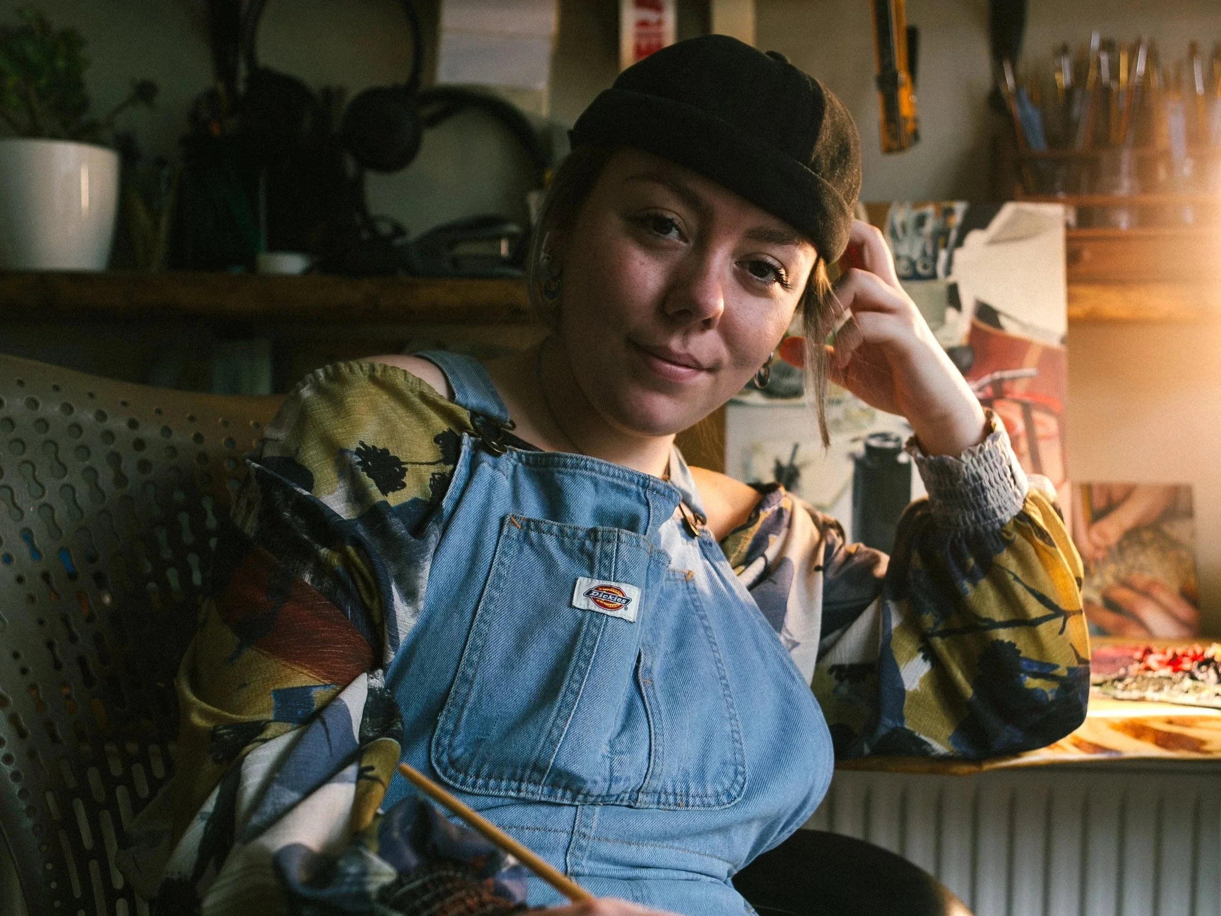 Young woman with short hair, wearing a black beret and colorful patterned shirt, sitting in a workshop or studio. She has a slight smile and is looking at the camera, with her hand resting on her head.