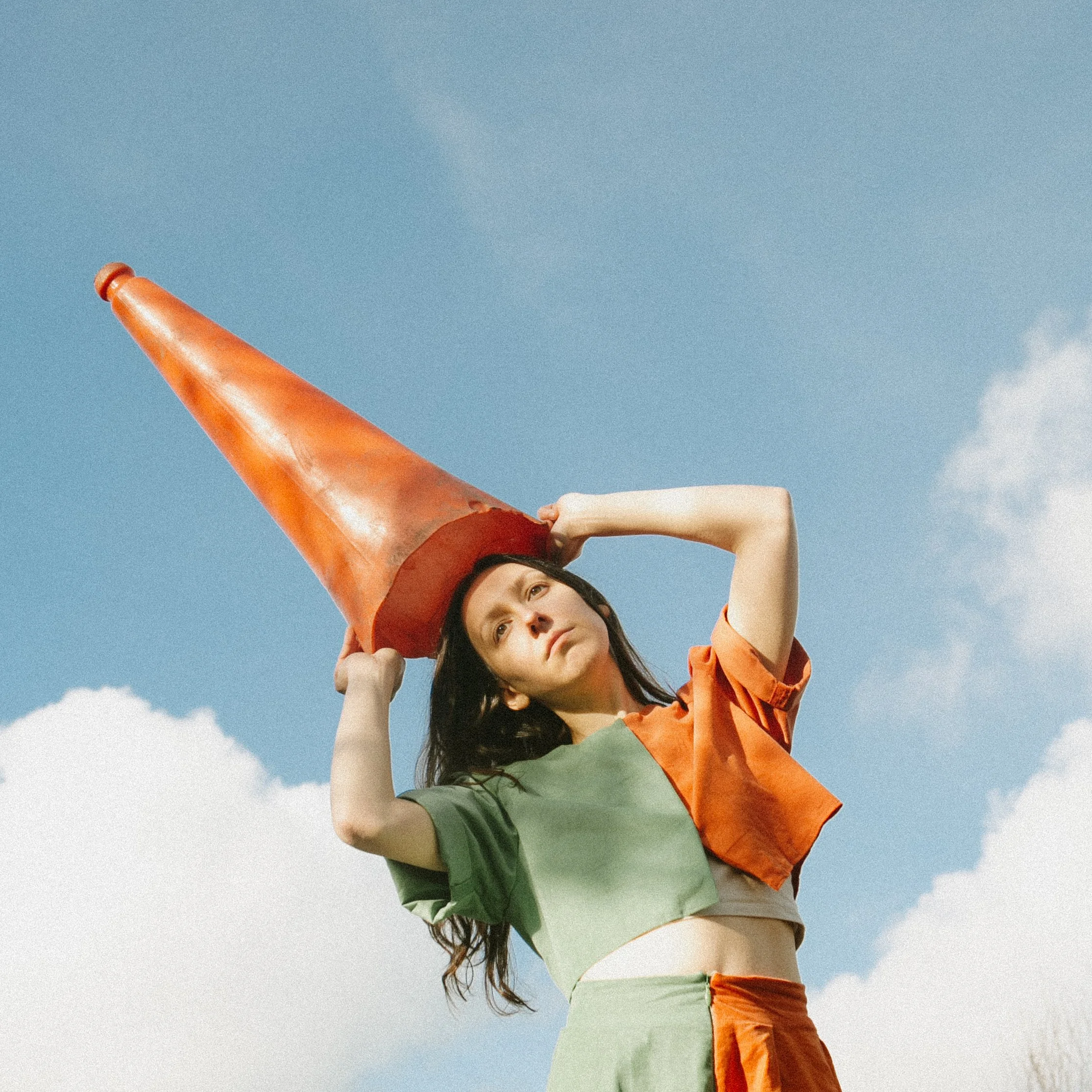 A woman standing outdoors against a blue sky with fluffy clouds, wearing an orange traffic cone on her head and a matching green and orange outfit.