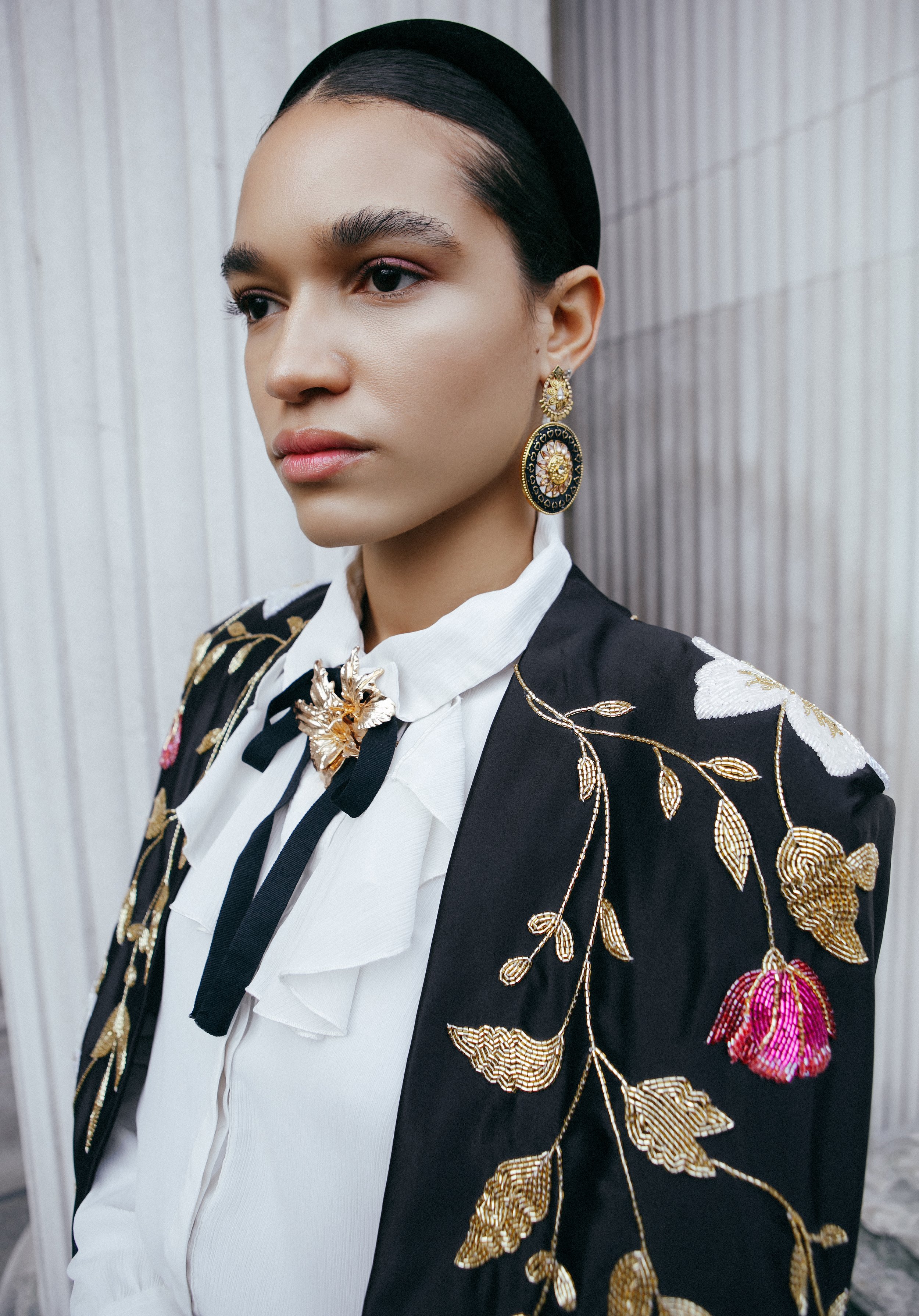 A woman wearing a black embroidered blazer with gold and pink floral details, a white blouse with a bow and a brooch, and large gold and black earrings. She has dark hair slicked back and is posing against a gray, vertically paneled background.