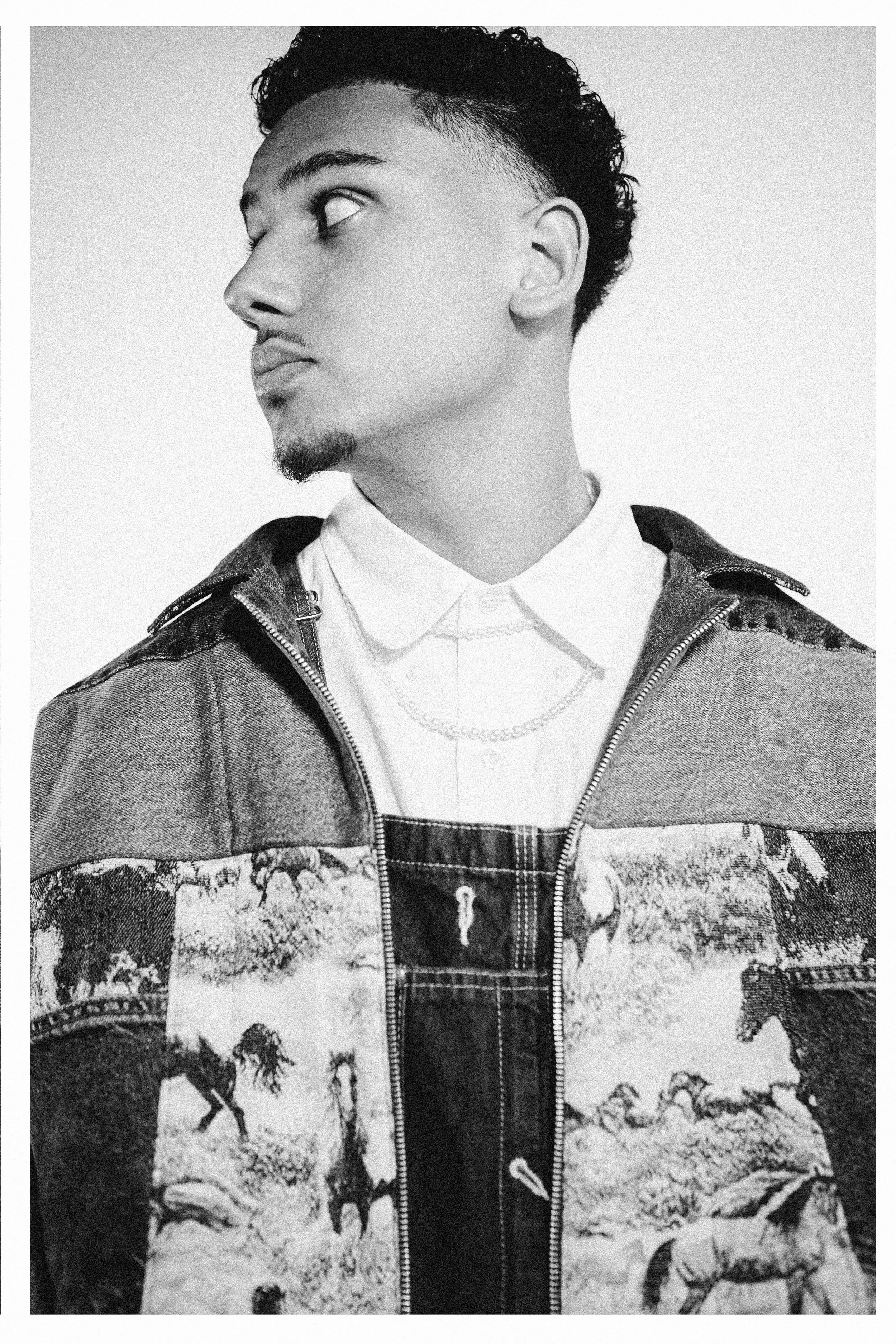 Black-and-white portrait of a young man with short, curly hair and a light mustache and goatee, looking to the side. He wears a patterned jacket over a collared shirt and layered necklaces.