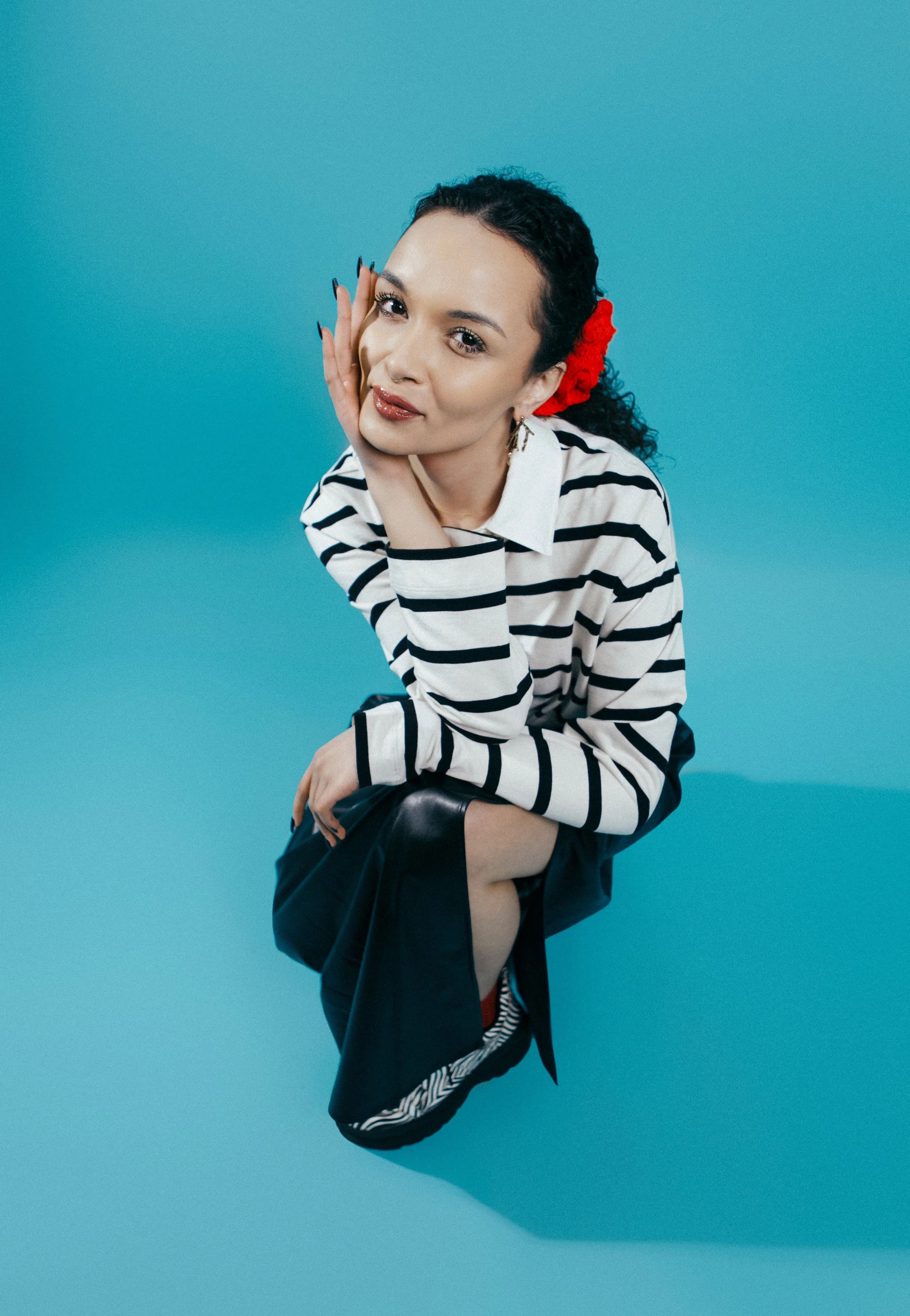 A woman with dark curly hair, wearing a striped black-and-white shirt and a red flower in her hair, kneeling on a blue background, resting her face on her hand and smiling softly.