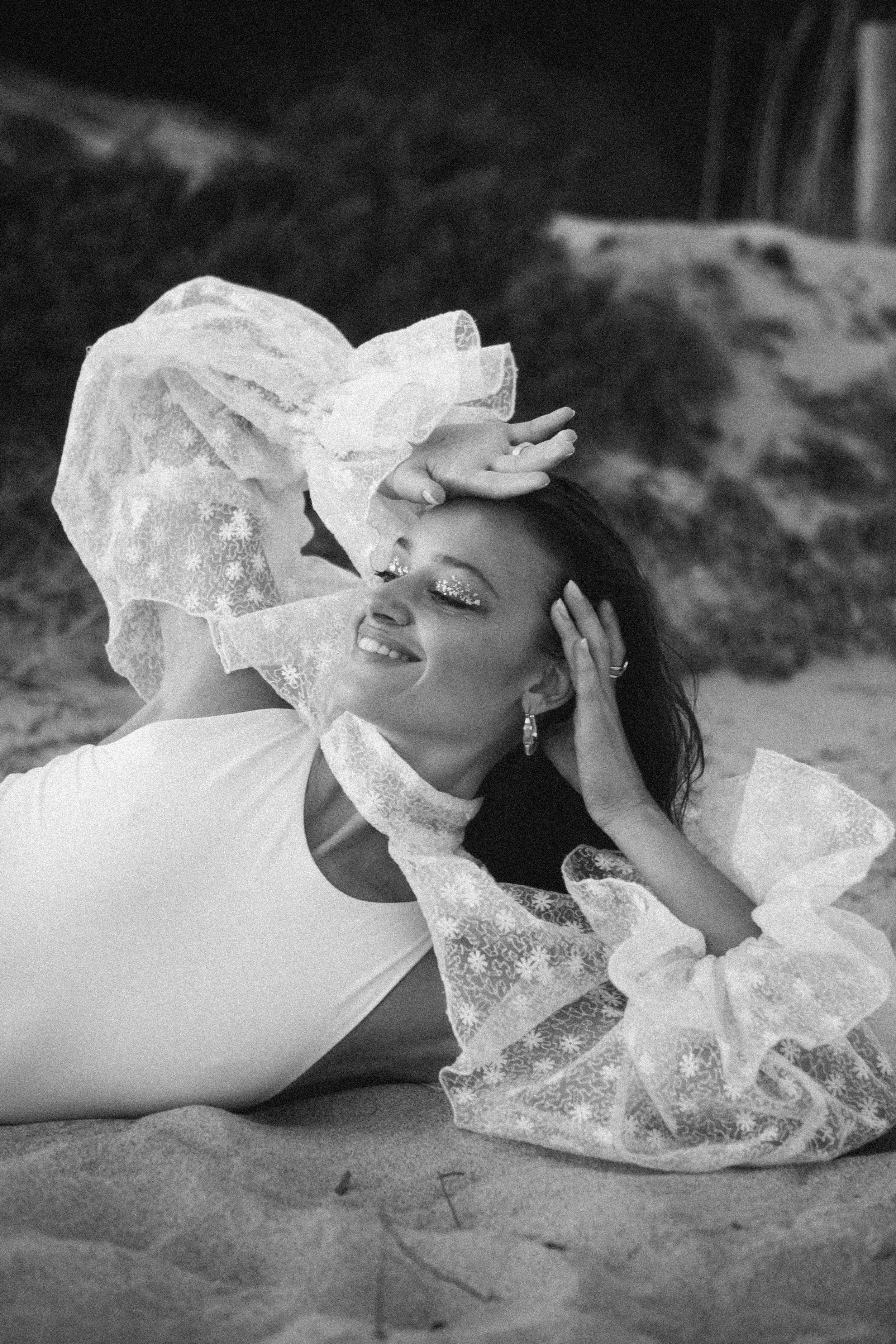 A woman lying on her side on a sandy surface, smiling with her eyes closed, wearing a white top and flared lace sleeves, with glitter on her eyelids and earrings, posing with one hand on her head and the other behind her head.