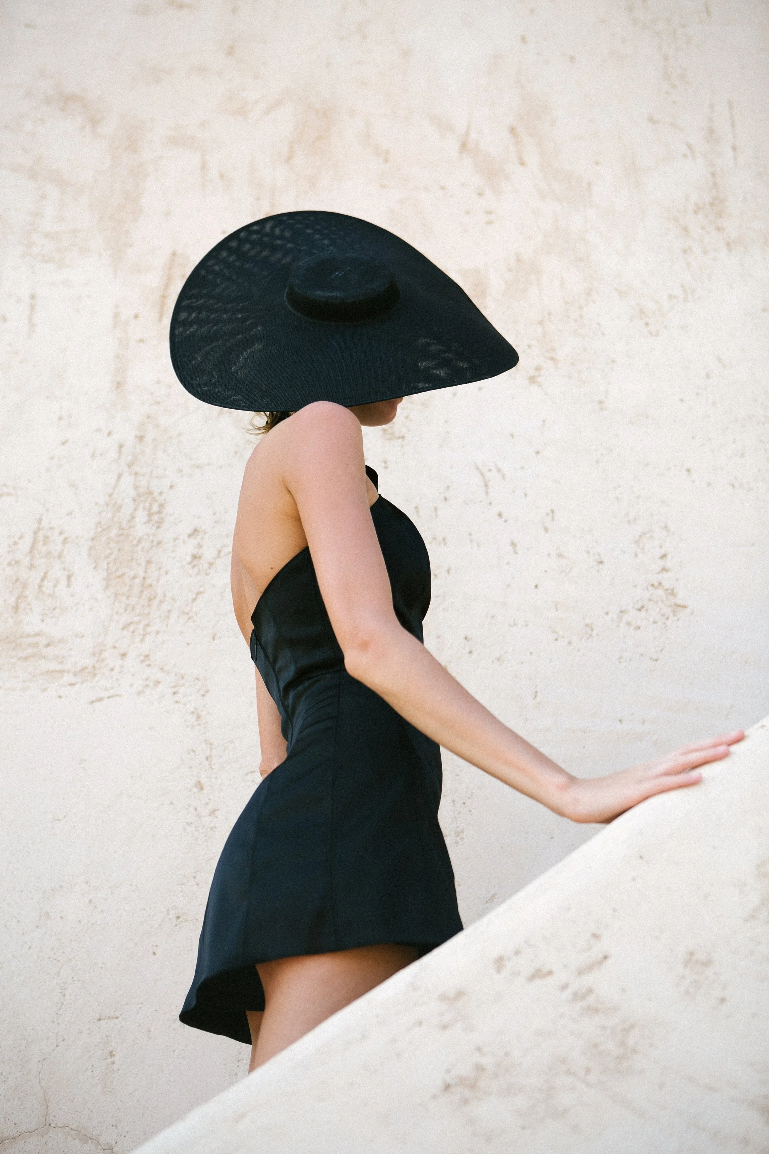 Woman in a black dress wearing a large black sunhat, leaning on a white surface against a textured light background.