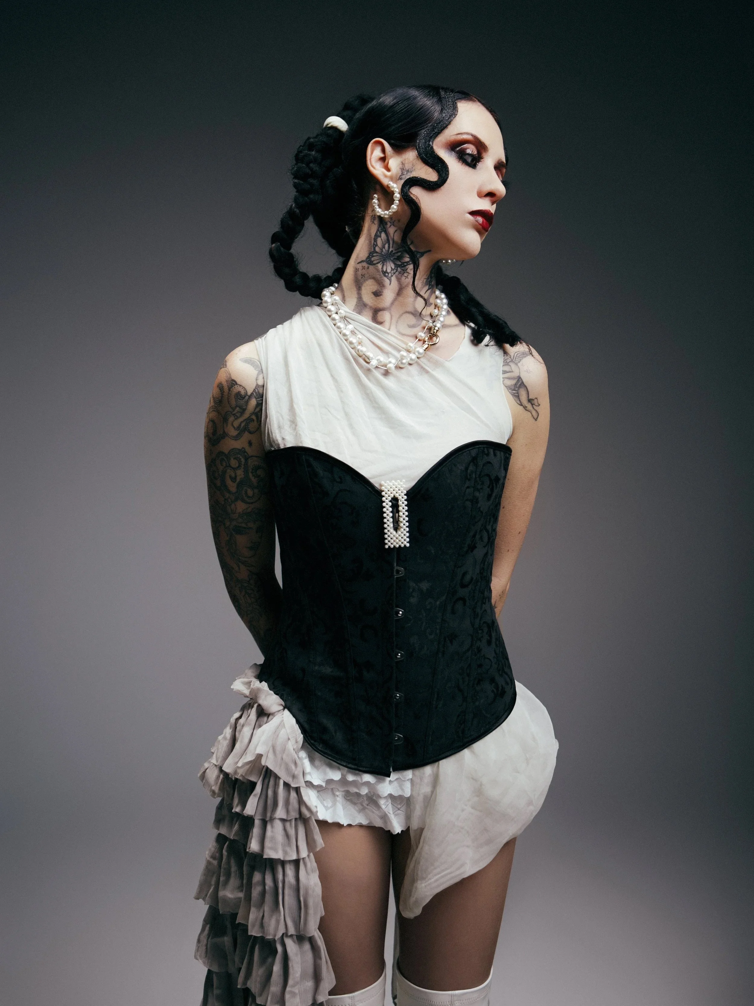 A woman with tattoos and dark hair styled in braids, wearing a black corset, ruffled skirt, and layered pearl necklaces, posing against a gray background.
