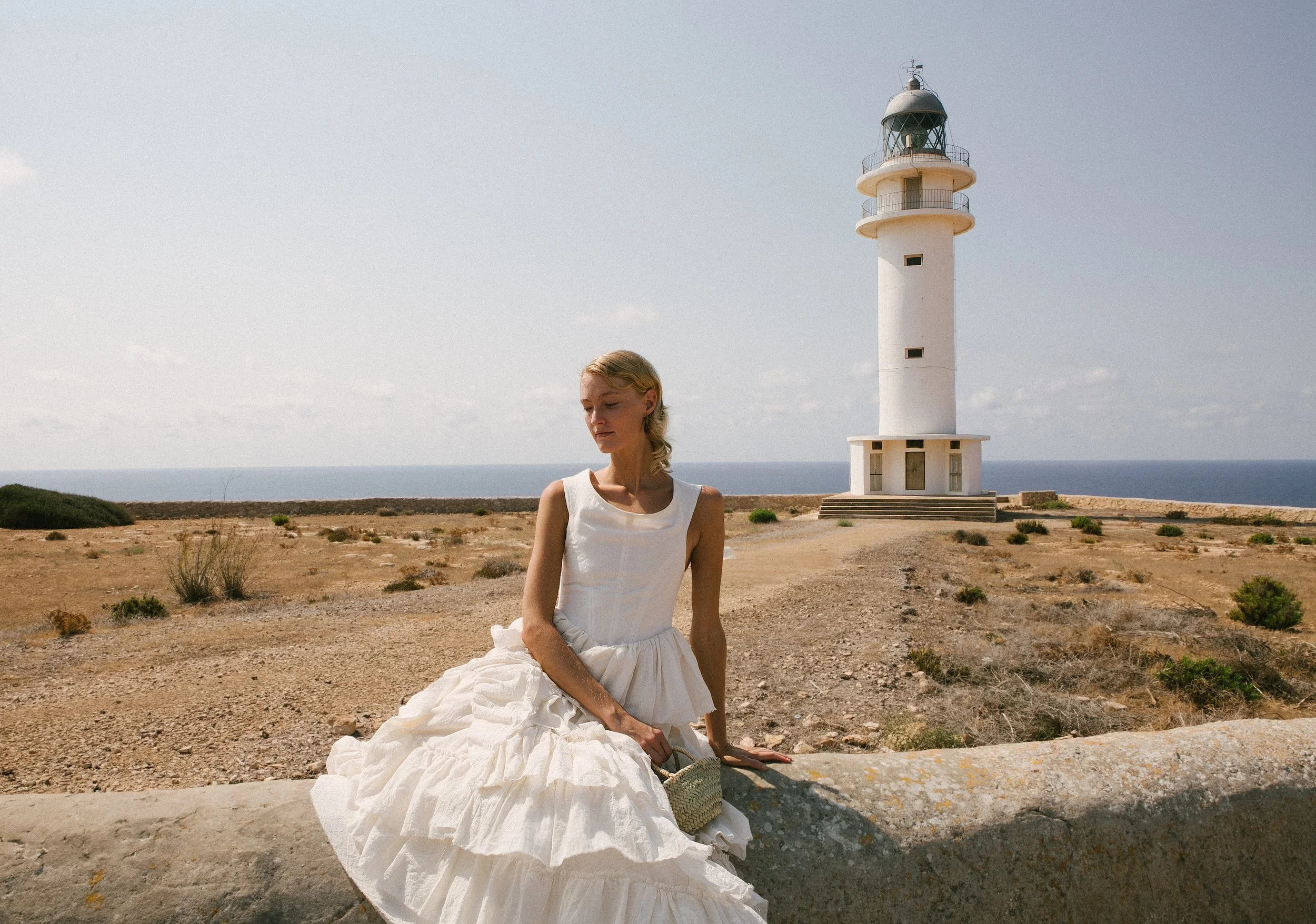 A woman in a white dress sitting on a stone wall near a lighthouse on a clear day with a view of the ocean in the background.