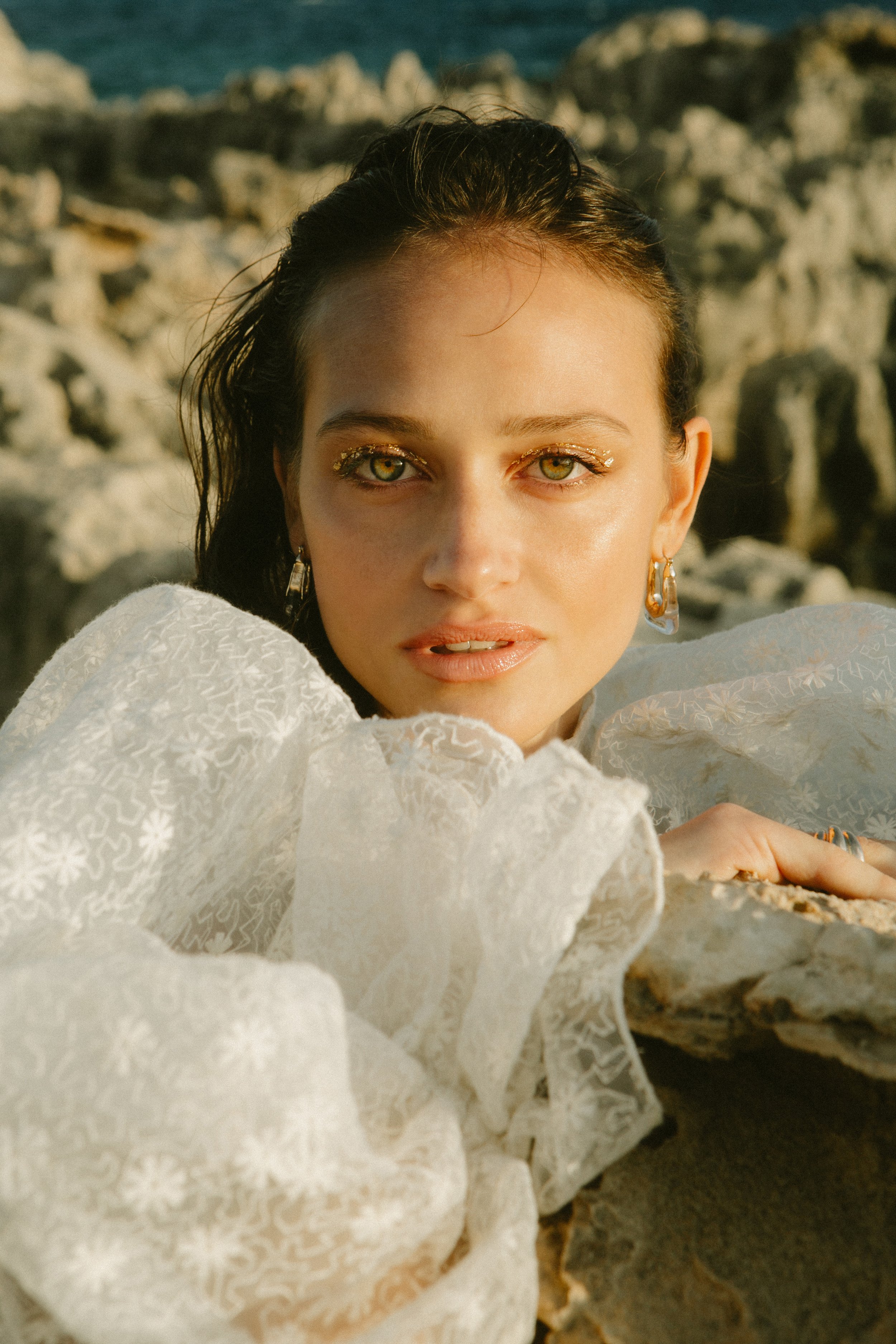 A young woman with golden eyes and gold glitter makeup lying on a rocky beach during sunset, wearing a white textured blouse and gold jewelry.