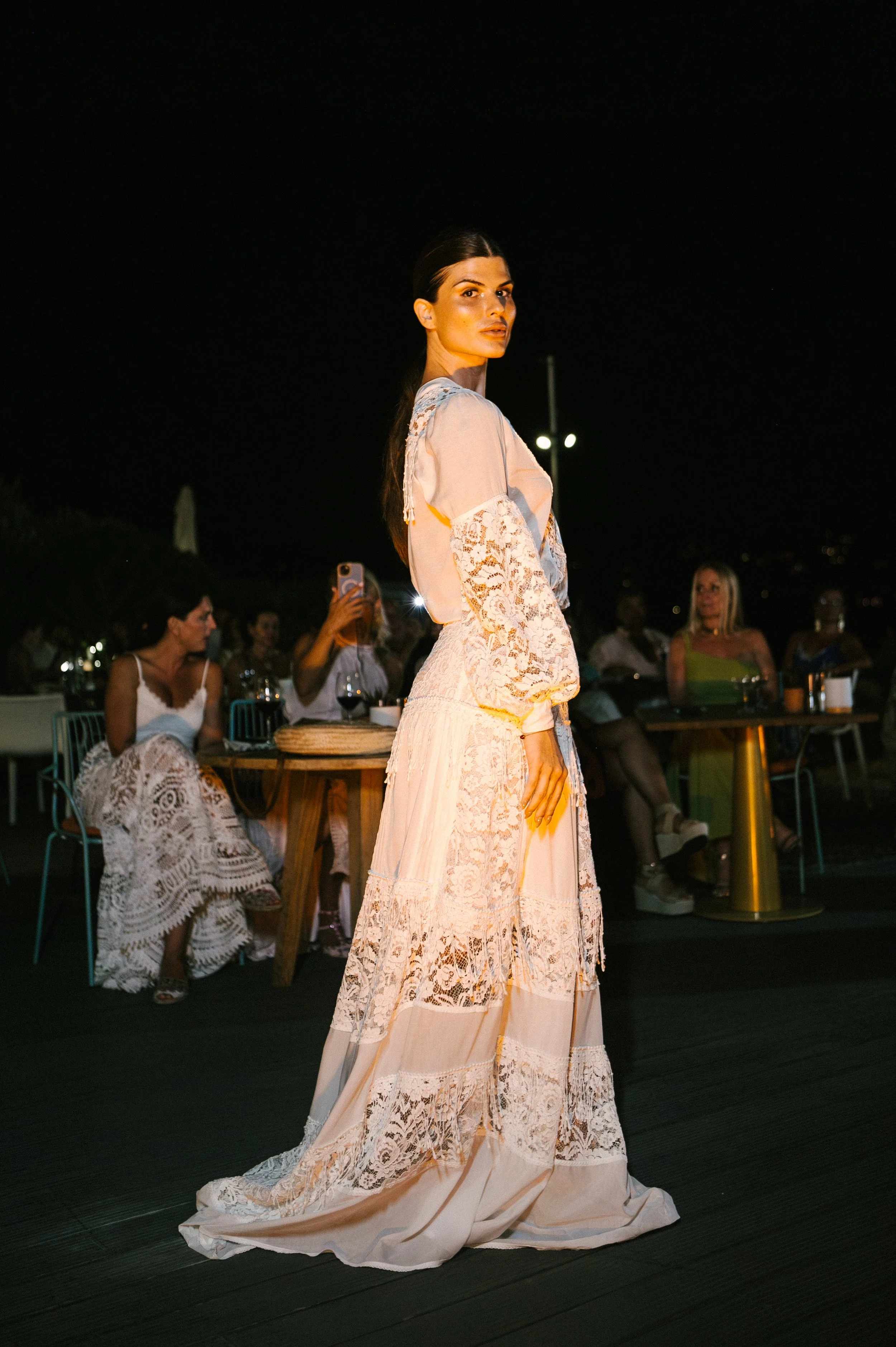A woman in a long, elegant lace dress standing outdoors at night while people sit at tables in the background.