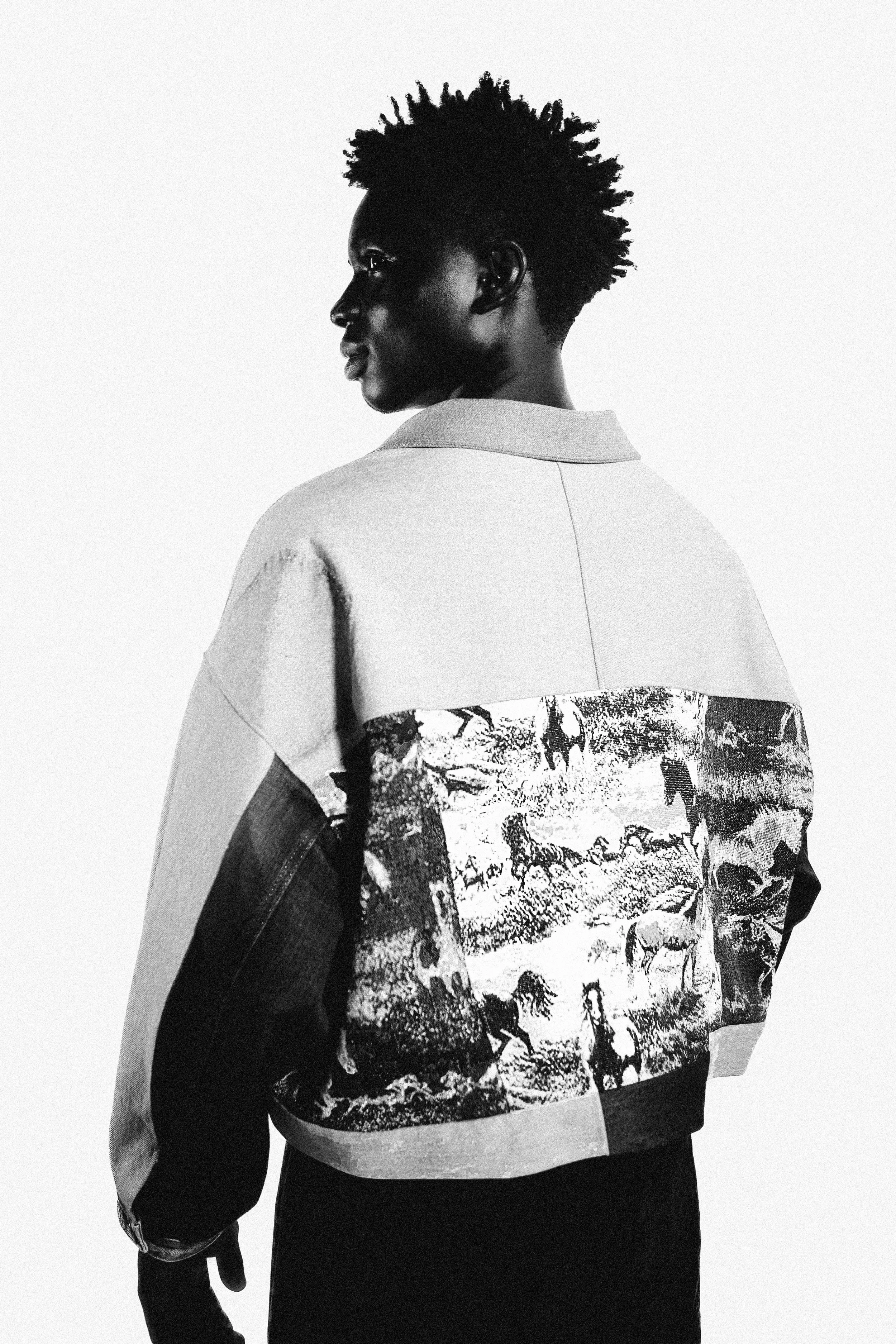 Black and white photo of a woman with short, spiky hair, looking to the left, wearing a jacket with an animal and landscape design on the back.