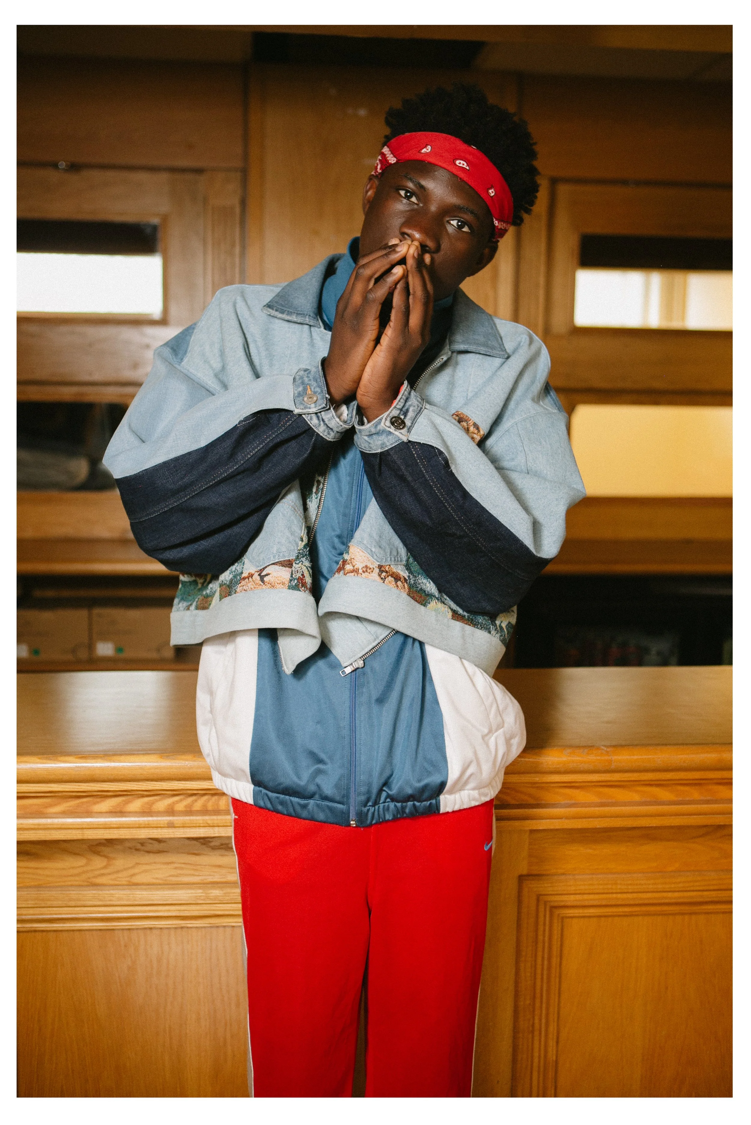 A young man with dark skin, wearing a red bandana, a vintage denim and patterned jacket, red Nike pants, and a blue zip-up shirt, standing indoors with a wooden background.
