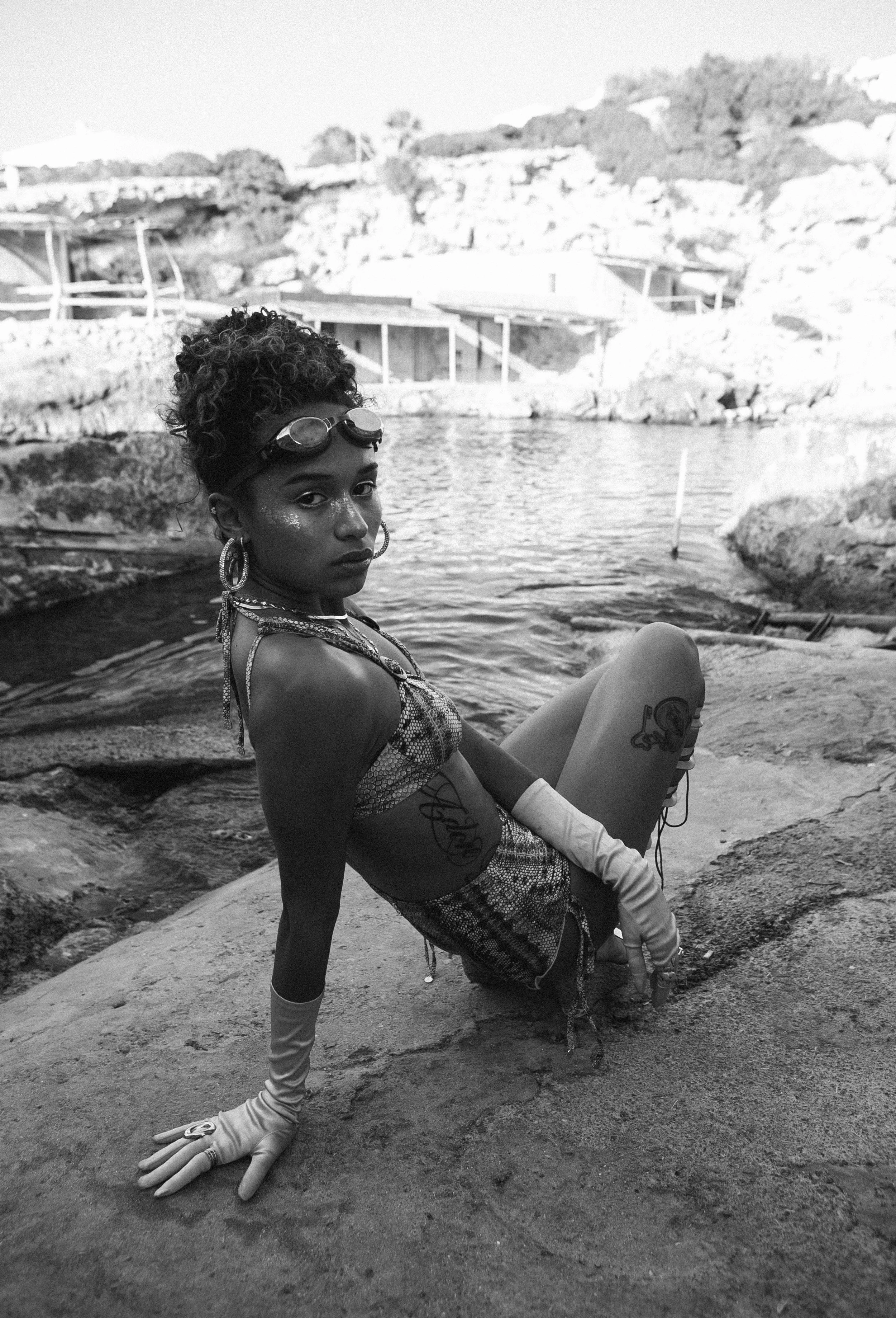 A young woman with curly hair, wearing goggles on her head, large hoop earrings, glitter makeup, and gloves, is sitting on a rock near water, with a rocky landscape in the background.