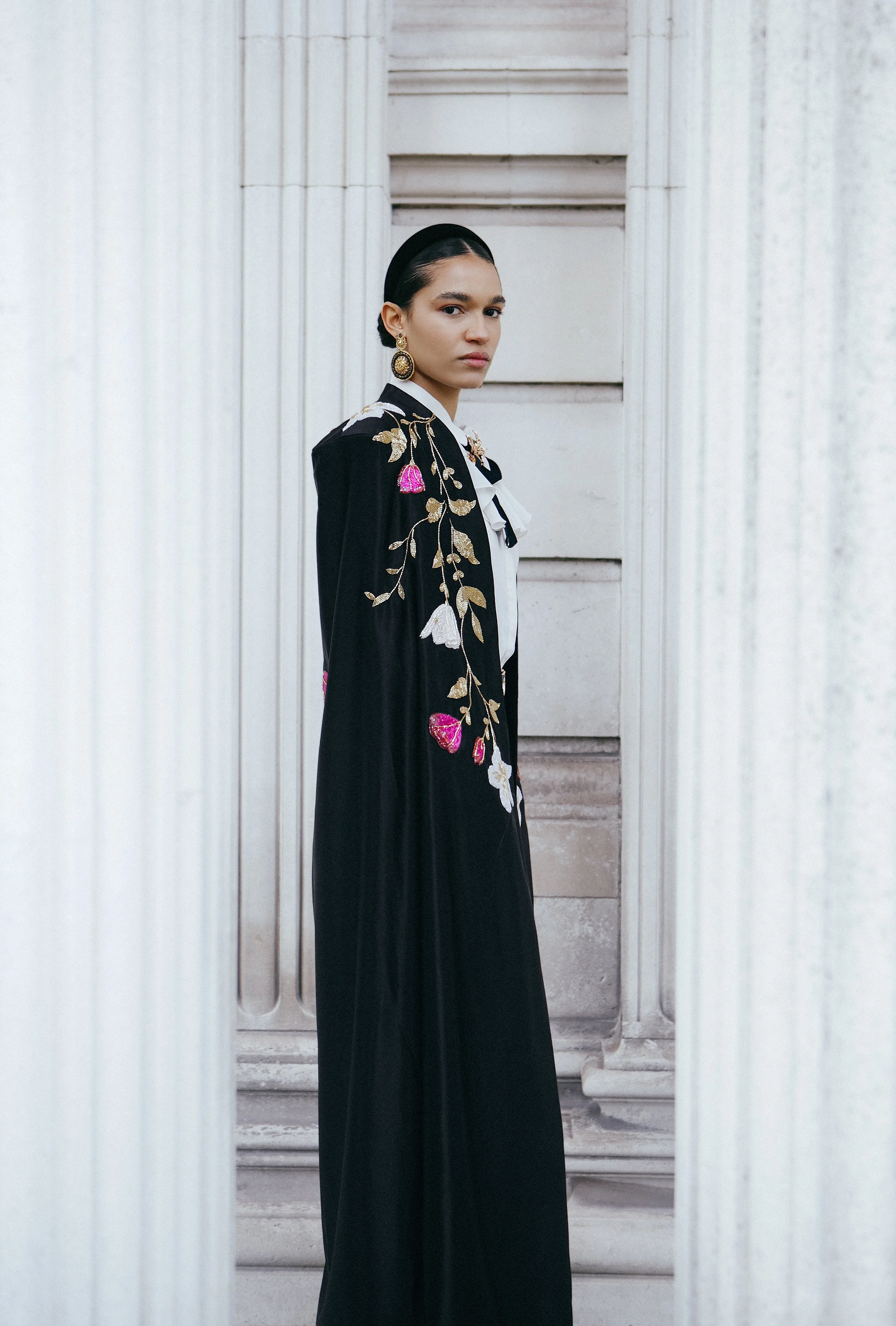 A woman standing between classical white columns, wearing a black embroidered cloak and earrings, with a serious expression.