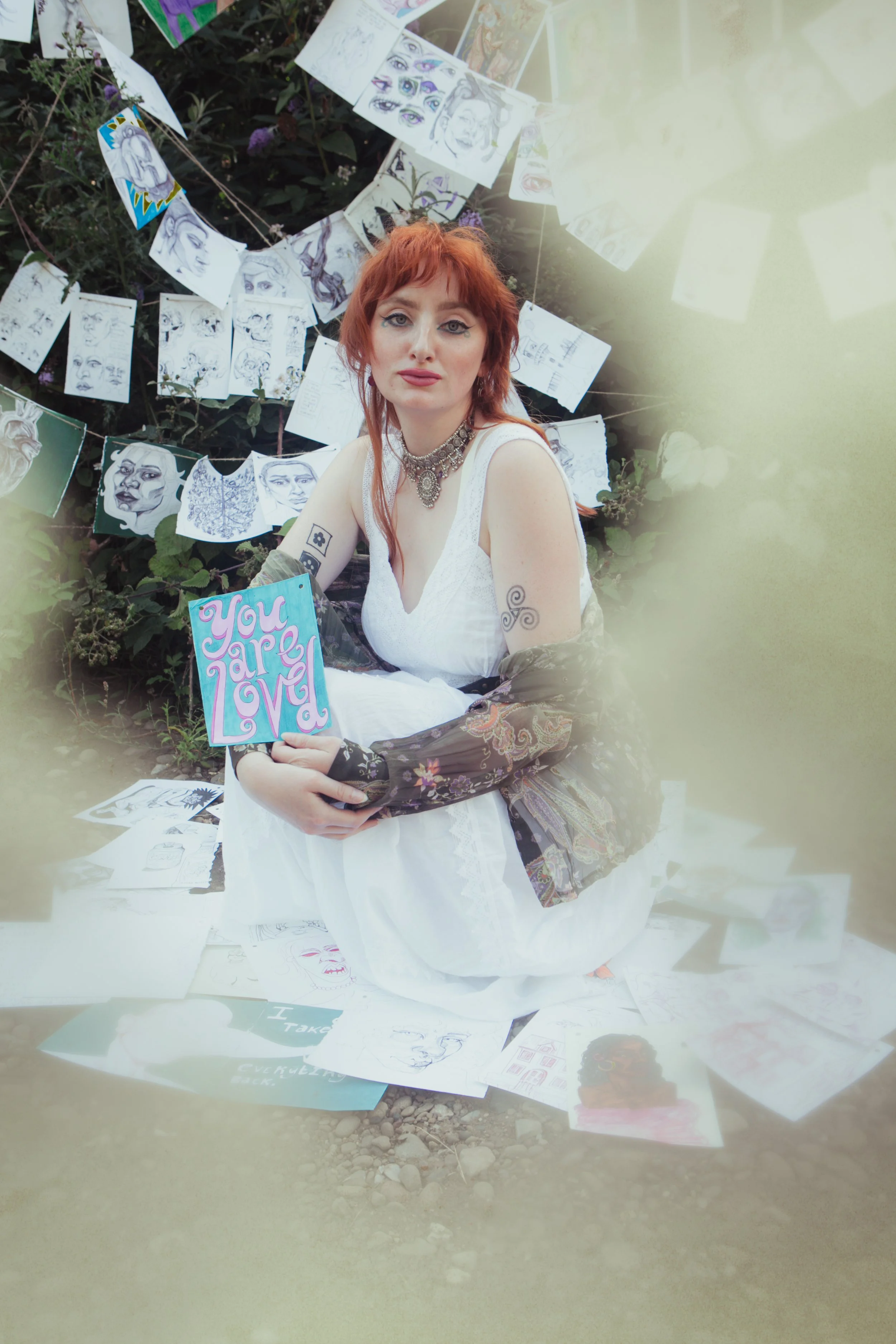 A woman with red hair and tattoos holding a colorful sign that says "You Are Loved," sitting outdoors surrounded by art sketches and paintings hanging on a string and scattered on the ground.