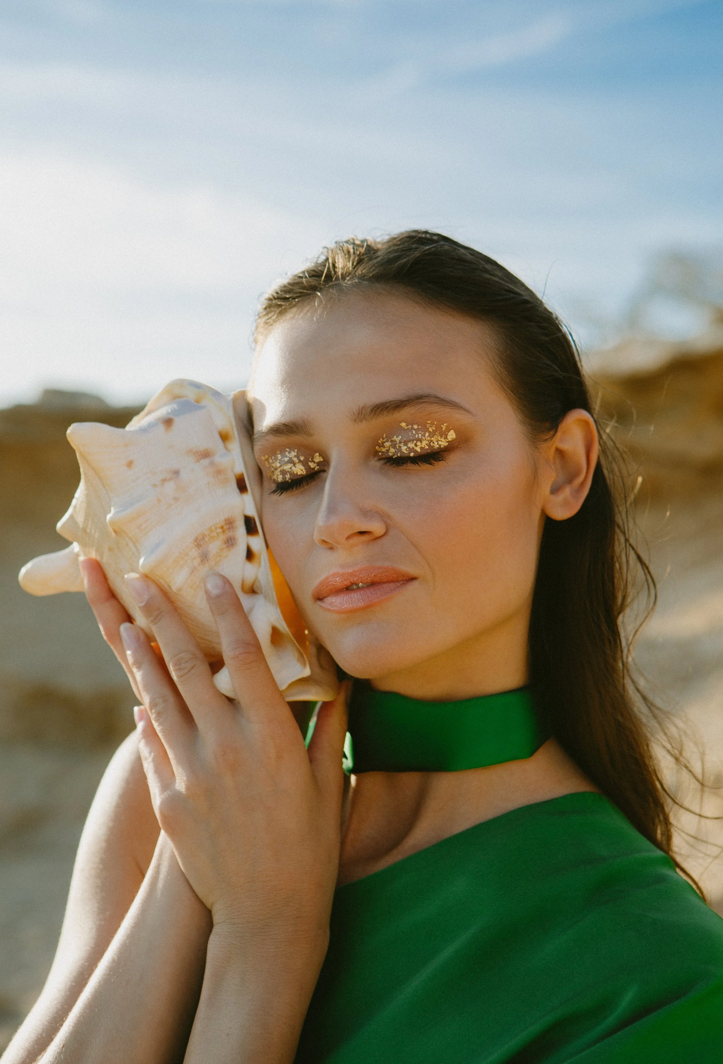 A woman with gold glitter makeup and a green dress holding a large seashell near her face outdoors.