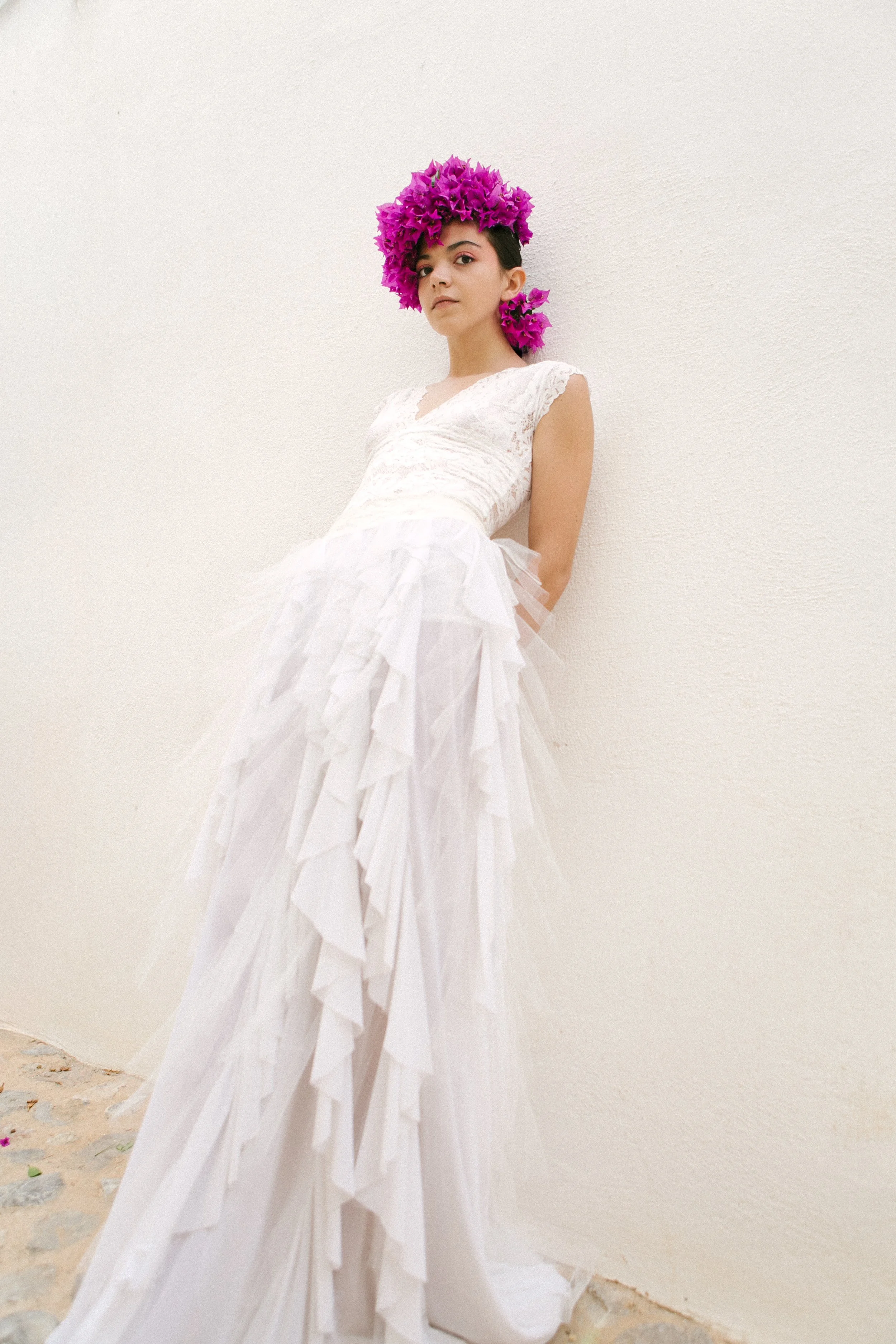 A woman wearing a white dress with ruffled layers and a crown of vibrant pink flowers standing against a plain cream wall.
