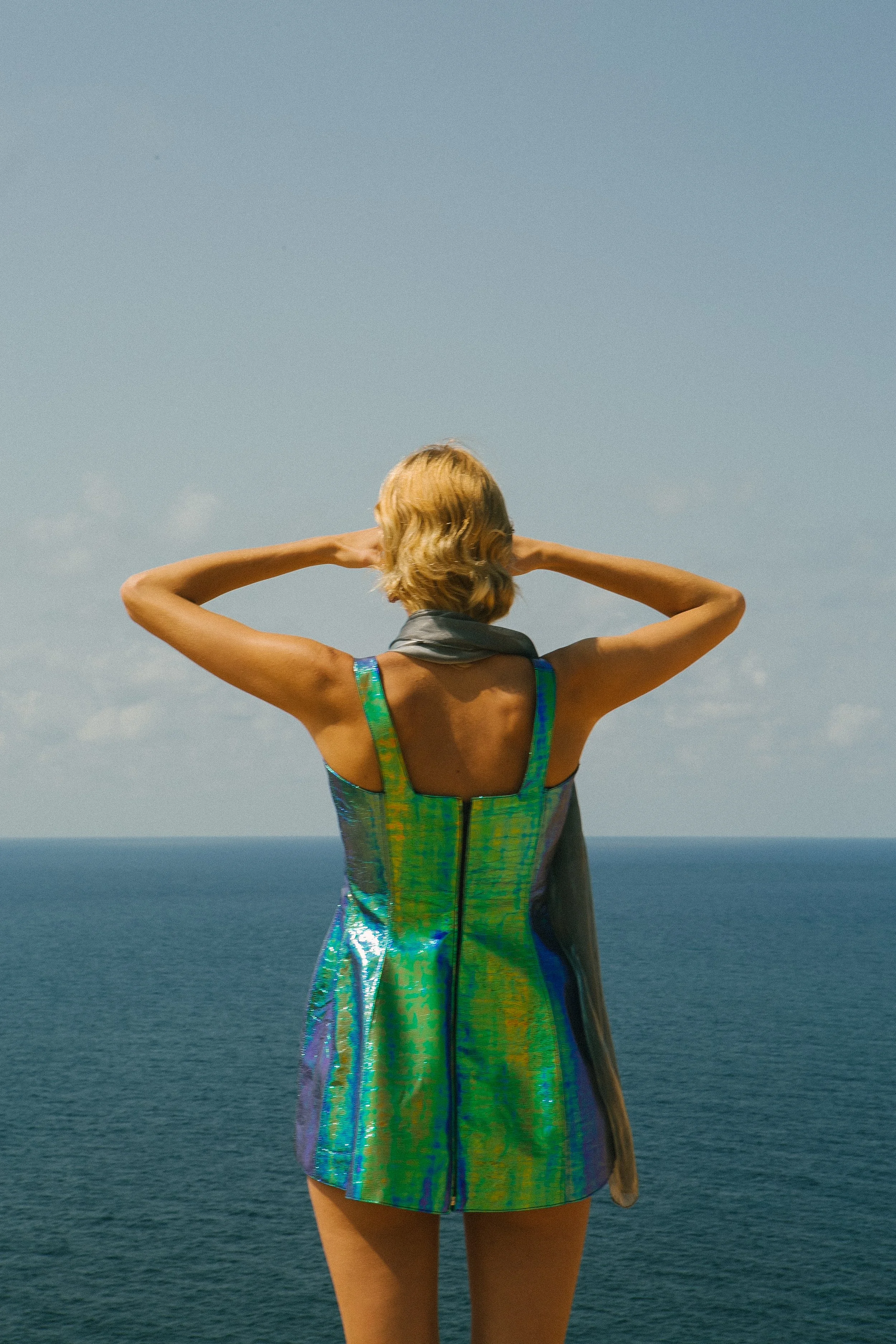 A woman with blonde hair standing with her back to the camera, looking out over the water with her hands behind her head. She's wearing a shiny, iridescent dress and a scarf around her neck.