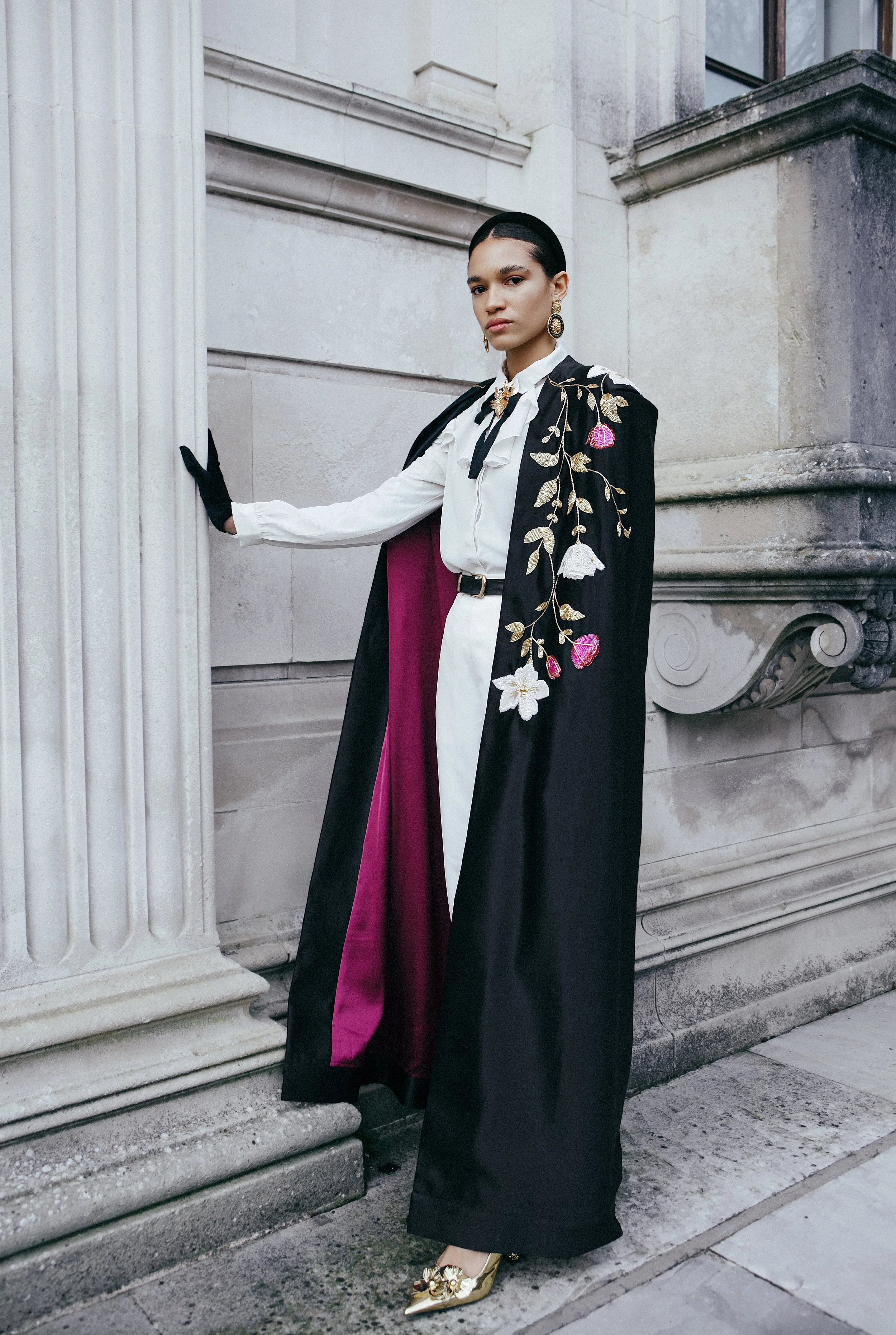 A woman dressed in a white blouse, black belt, and black wide-leg pants, wearing a black cape with floral embroidery and a purple lining, standing against a stone building with classical architectural details.