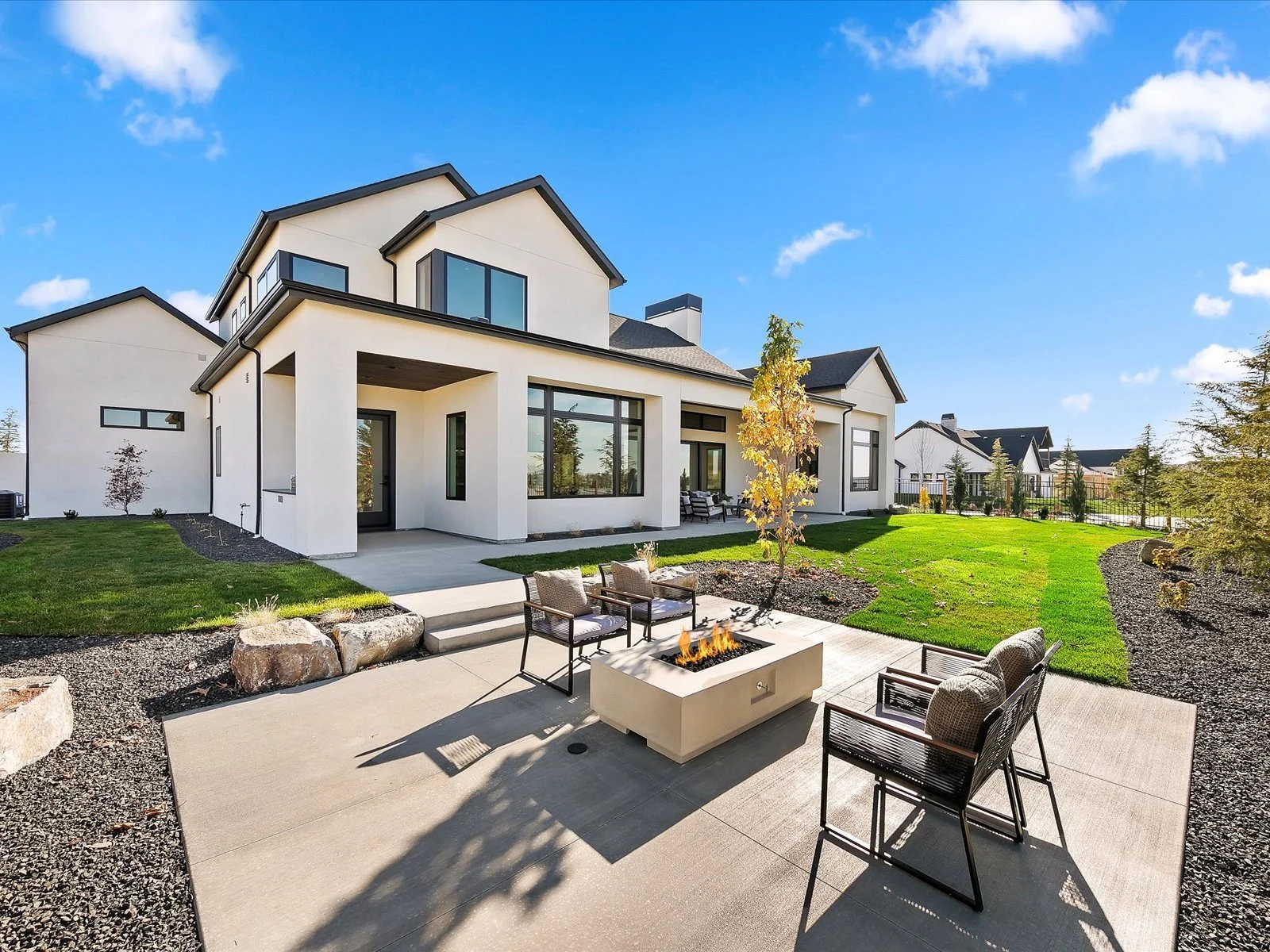 Modern white house with large windows, a landscaped yard, a patio area with outdoor seating and a fire feature, under a bright blue sky with a few clouds.