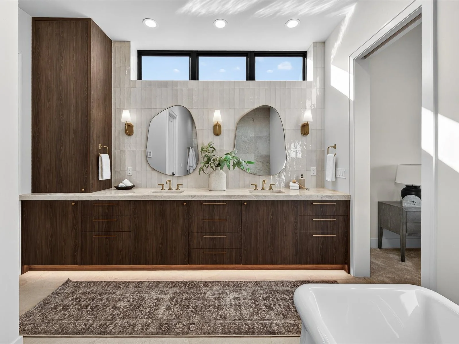 Modern bathroom with double vanity, dark wood cabinets, marble countertop, two oval mirrors, gold fixtures, wall sconces, a large window, a bathtub in the foreground, and a small side table with a lamp in the background.