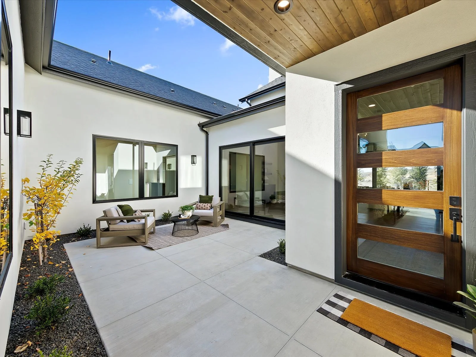 Modern outdoor patio with two cushioned chairs and a small round table, next to a white house with large sliding glass doors and a wooden front door, under a wooden ceiling.