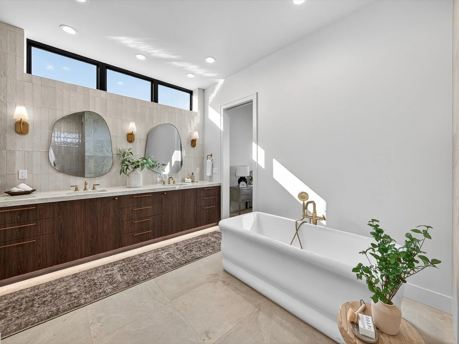 Modern bathroom with a double vanity featuring a white countertop, two round mirrors, and three wall sconces. A white bathtub with gold fixtures is beside a small wooden table holding a plant in a beige pot. Large window bringing in natural light is 