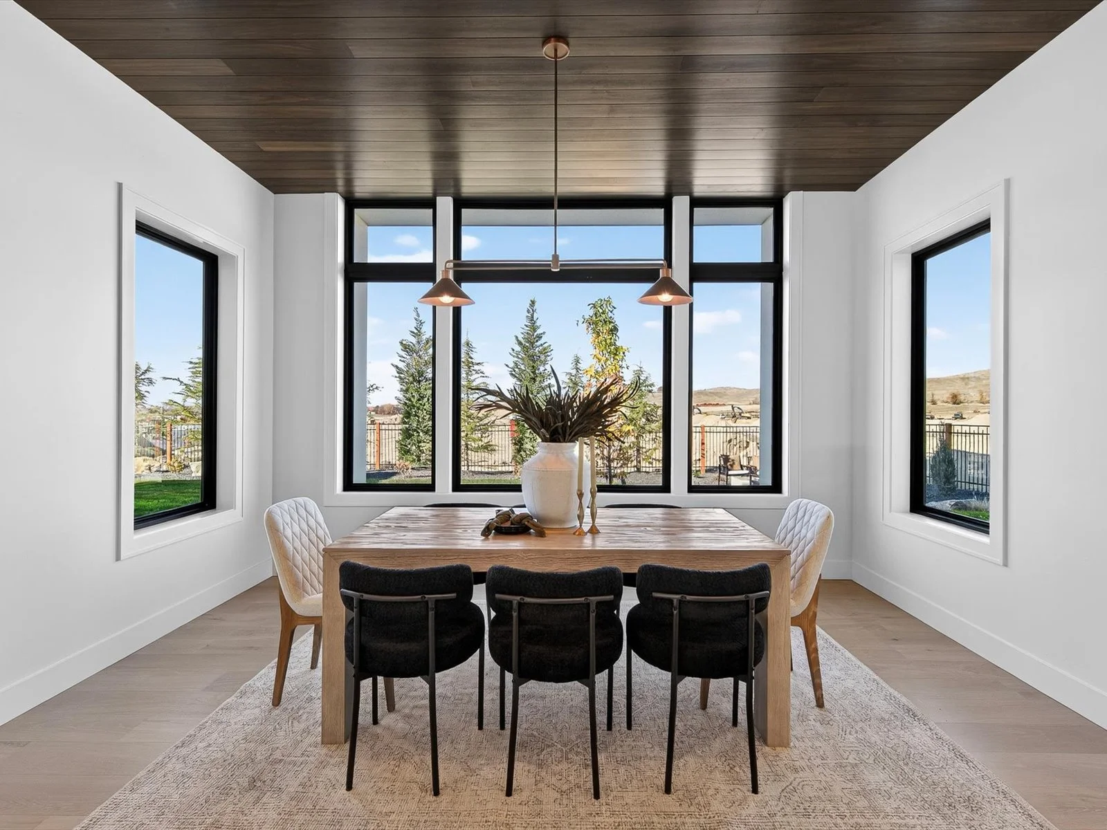 Modern dining room with a wooden table, six black and white upholstered chairs, large window with a view of trees and a fence, and a beige area rug.