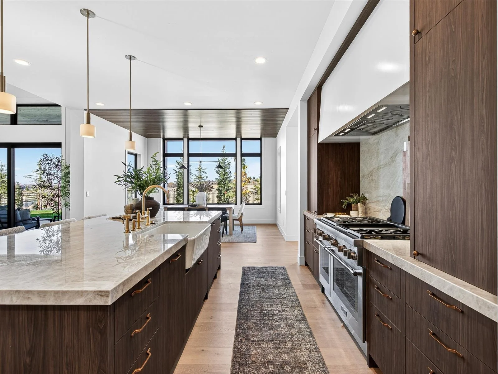 Modern kitchen with dark wood cabinets, stainless steel stove, white marble island with a sink, pendant lights, and large windows showing a garden outside.