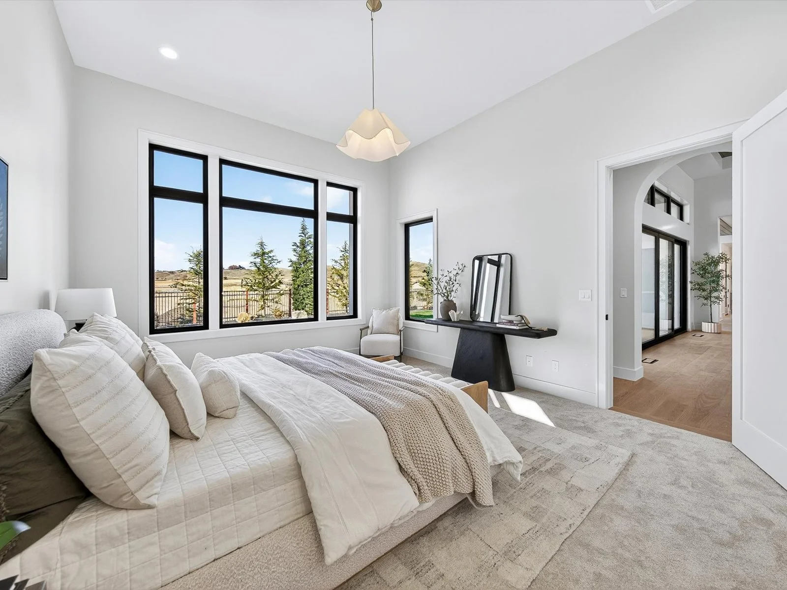 A modern bedroom with a large bed, multiple pillows, a nightstand with a lamp, and a large window with a view of trees and open land. There is a small chair, a black console table with a mirror and decorative items, and an adjoining room with sliding