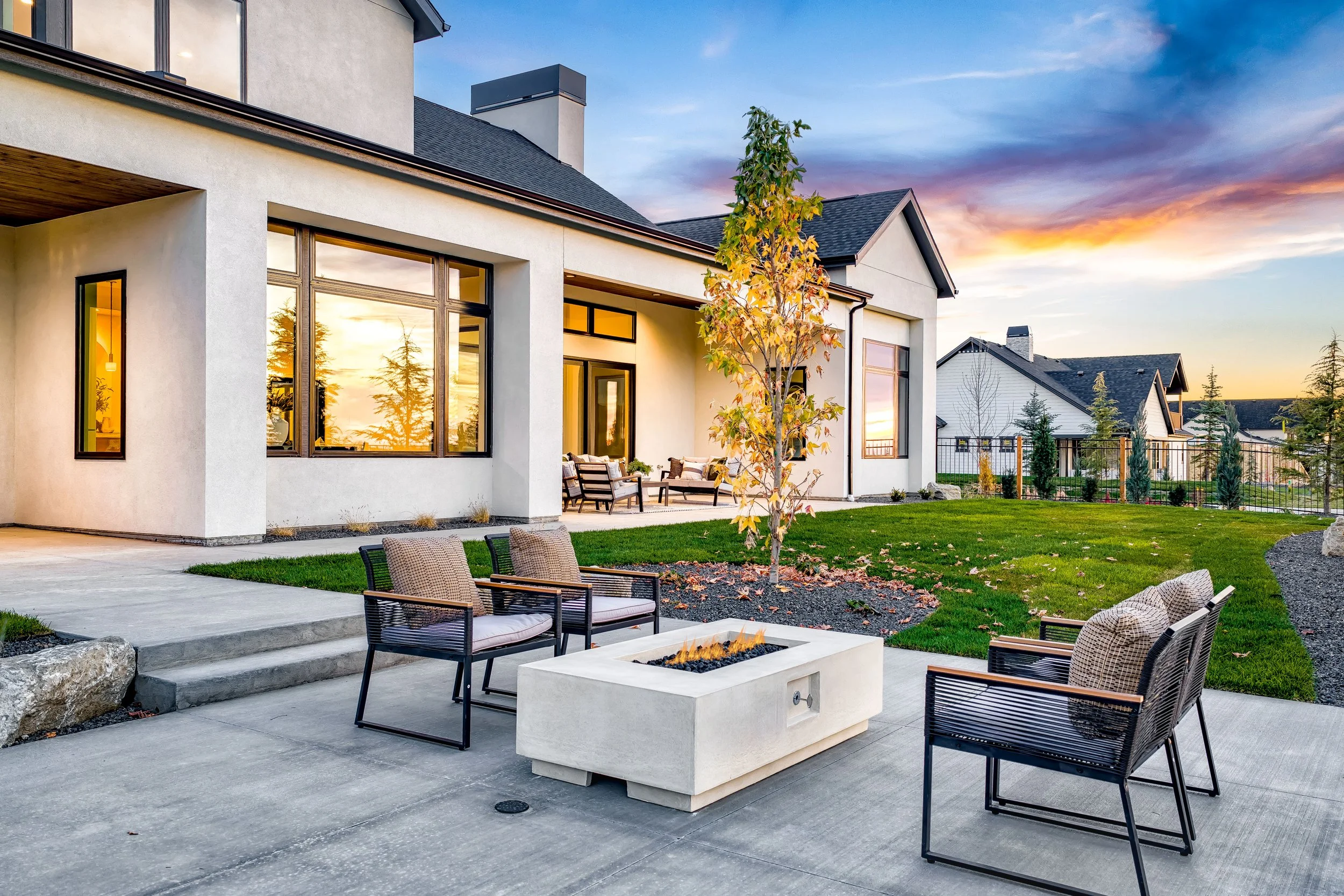 A modern backyard patio with a fire pit, four chairs, a lush green lawn, and a house with large windows reflecting the sunset sky.