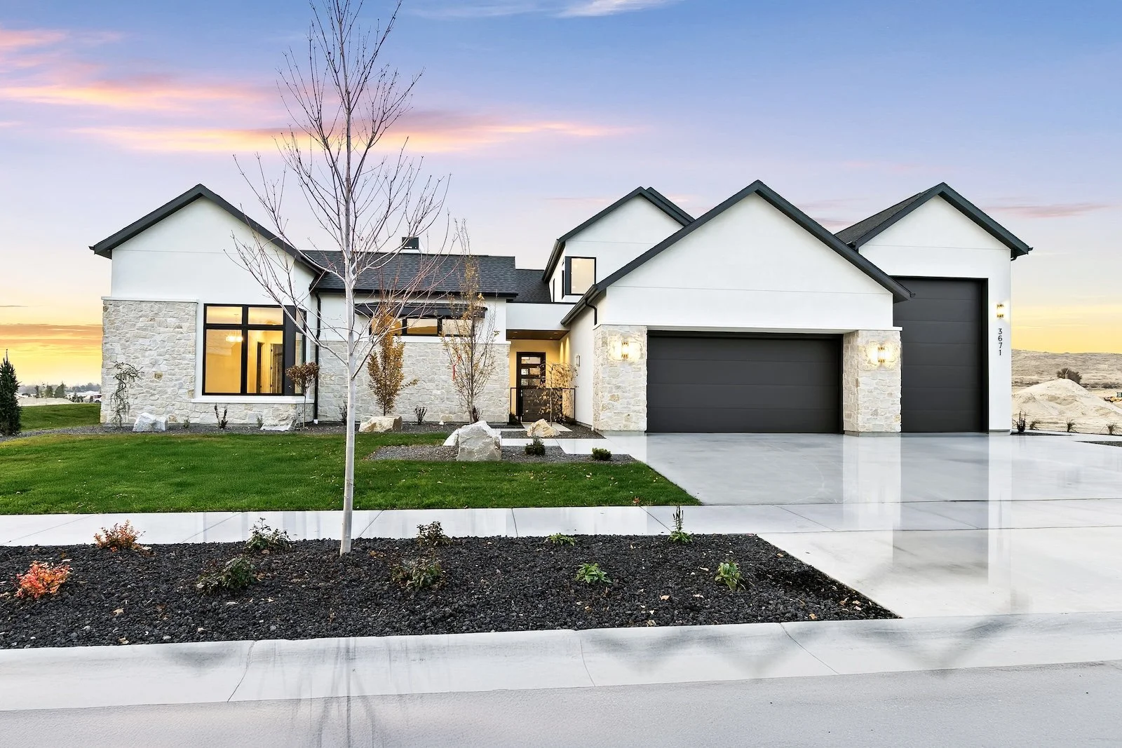 Modern two-story house with light-colored stone and white siding exterior, black garage doors, and large windows, during sunset with a landscaped front yard and driveway.
