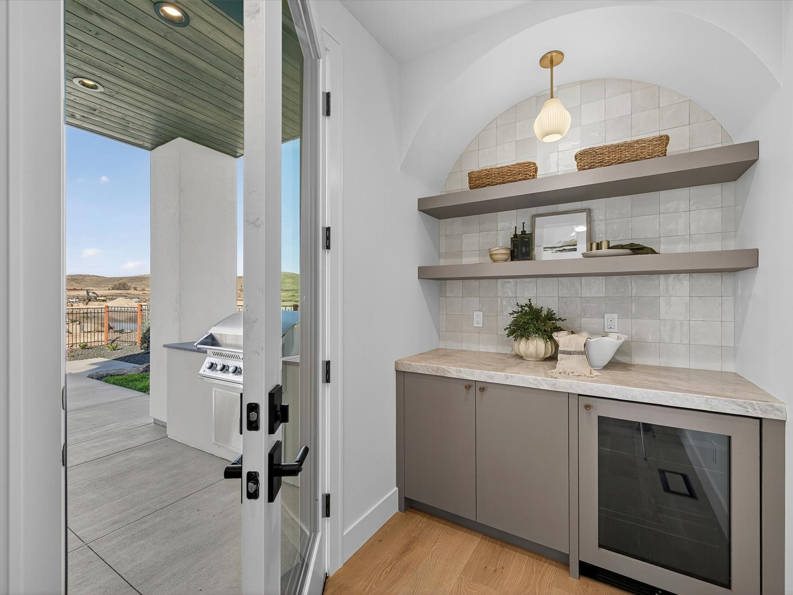 Interior of a room with a sliding glass door leading to an outdoor patio with a barbecue grill, open sky, and hills in the background. Inside, there are two floating shelves with woven baskets, decorative items, and a mirror. A countertop with a pott