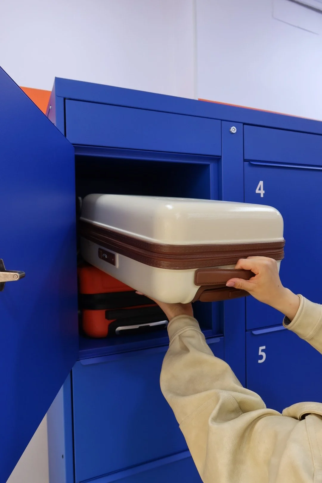 Person placing a cream-colored hard shell suitcase into a blue locker numbered 4.